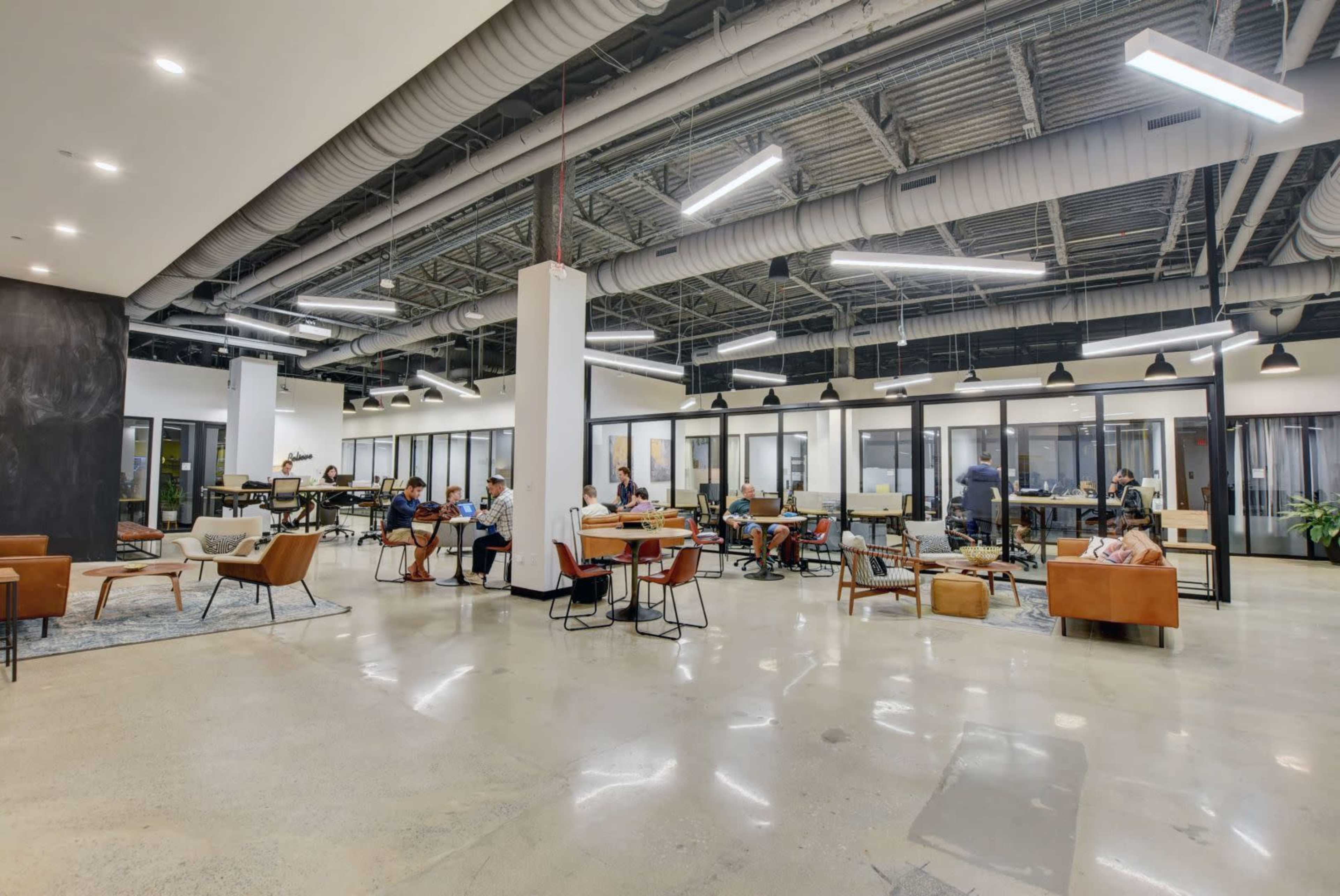 A modern office space with several workstations, lounge areas, and glass-enclosed meeting rooms, populated by individuals engaged in various activities.