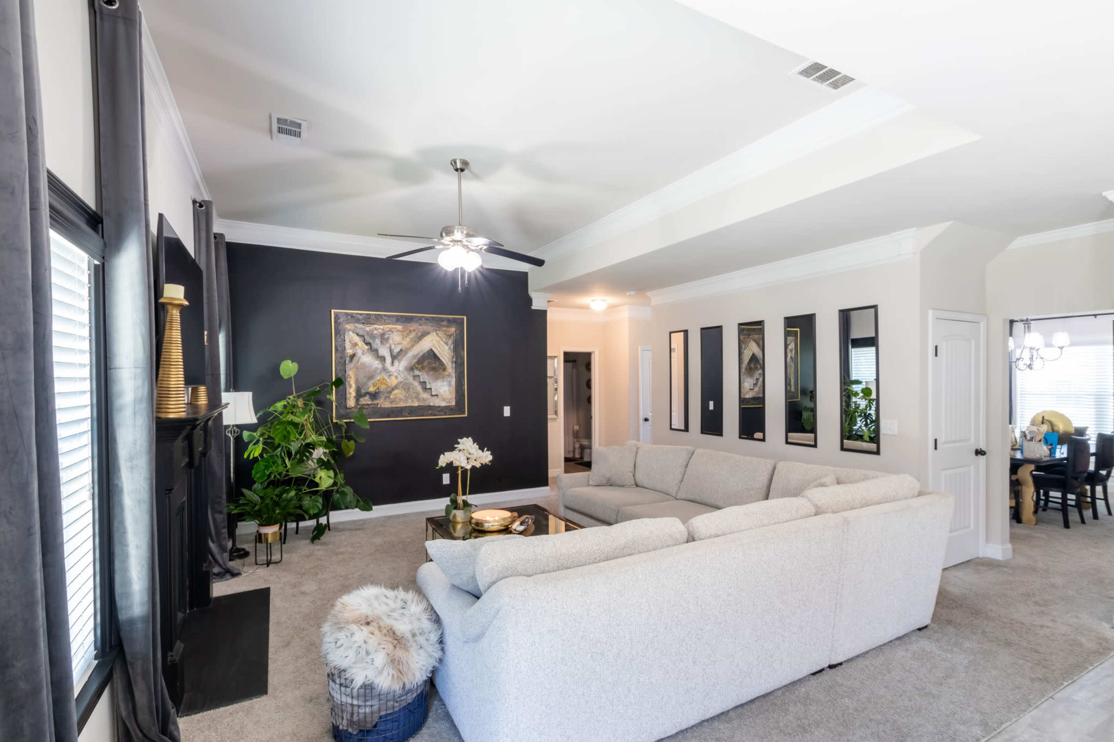 The image shows a cozy living room with a large light-colored sectional sofa, a dark accent wall featuring artwork, and various plants decoratively placed around the space.
