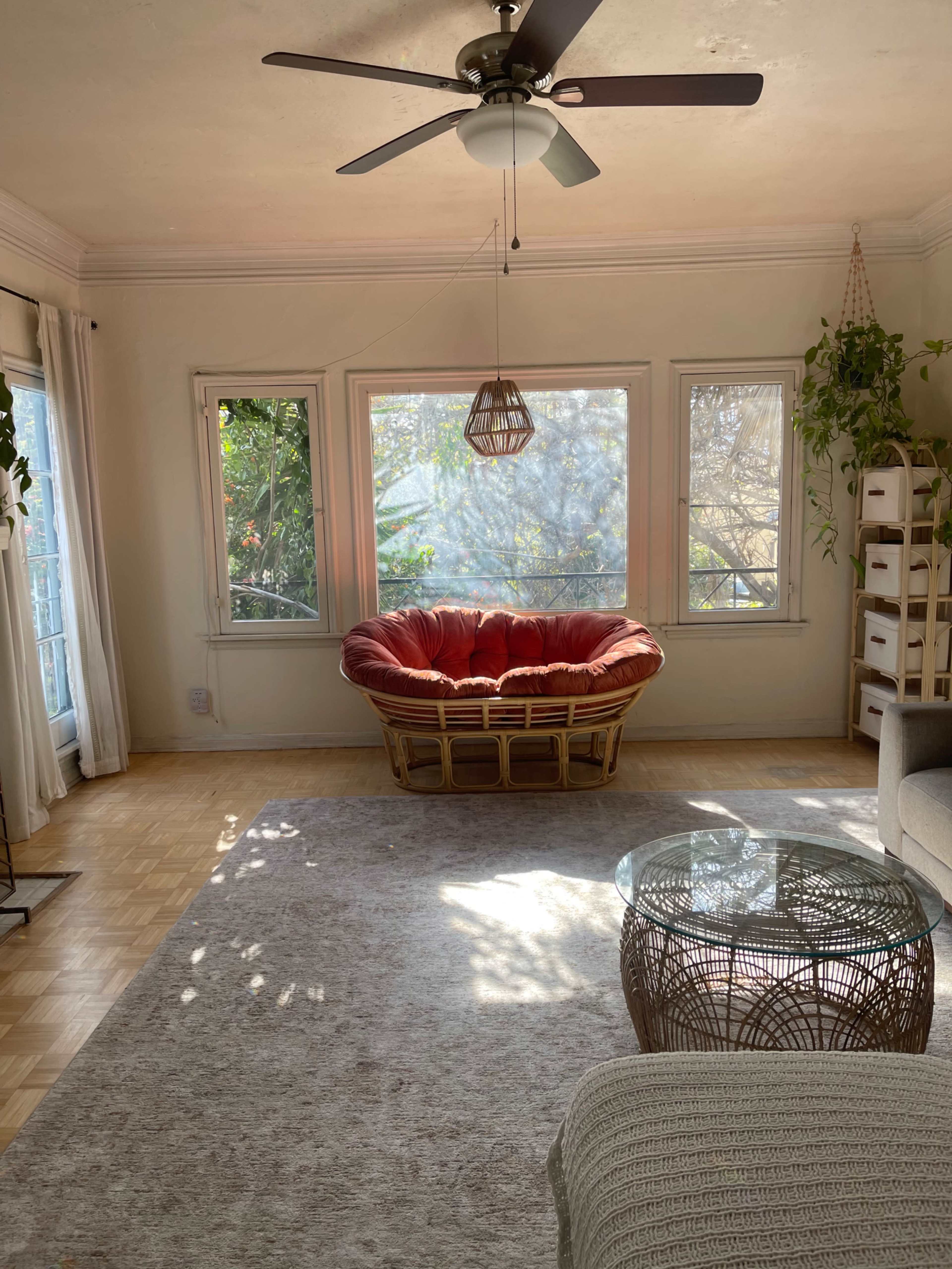The image shows a bright living room featuring a large circular wicker chair, a glass coffee table, and several windows that allow natural light to enter the space.