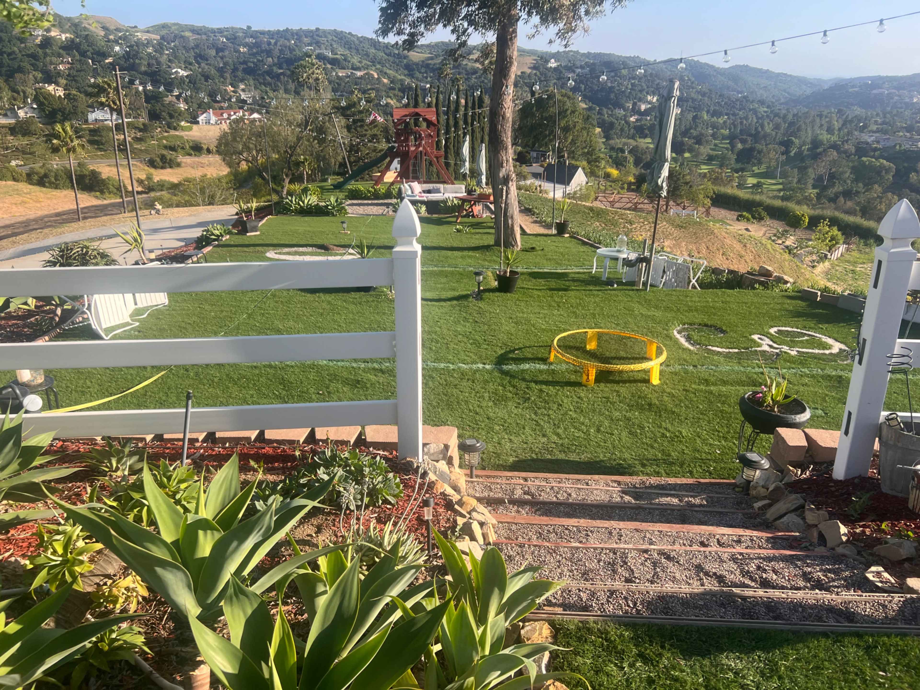 A landscaped outdoor area with green grass, stone pathways, and a play structure in the background, set against a mountainous landscape.