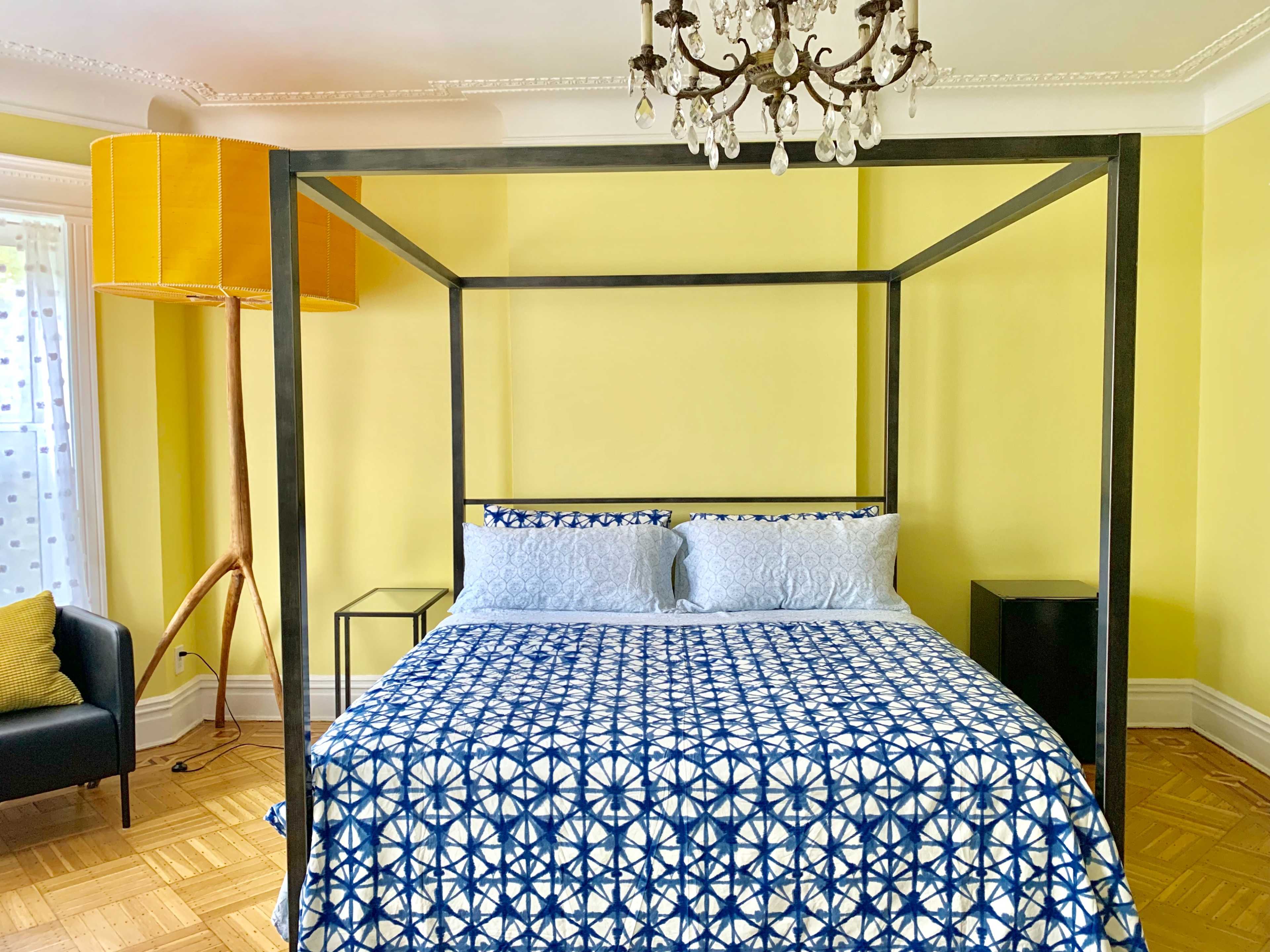 A bedroom with a four-poster bed covered in a blue and white patterned duvet, accompanied by a black armchair and a tall yellow lamp.