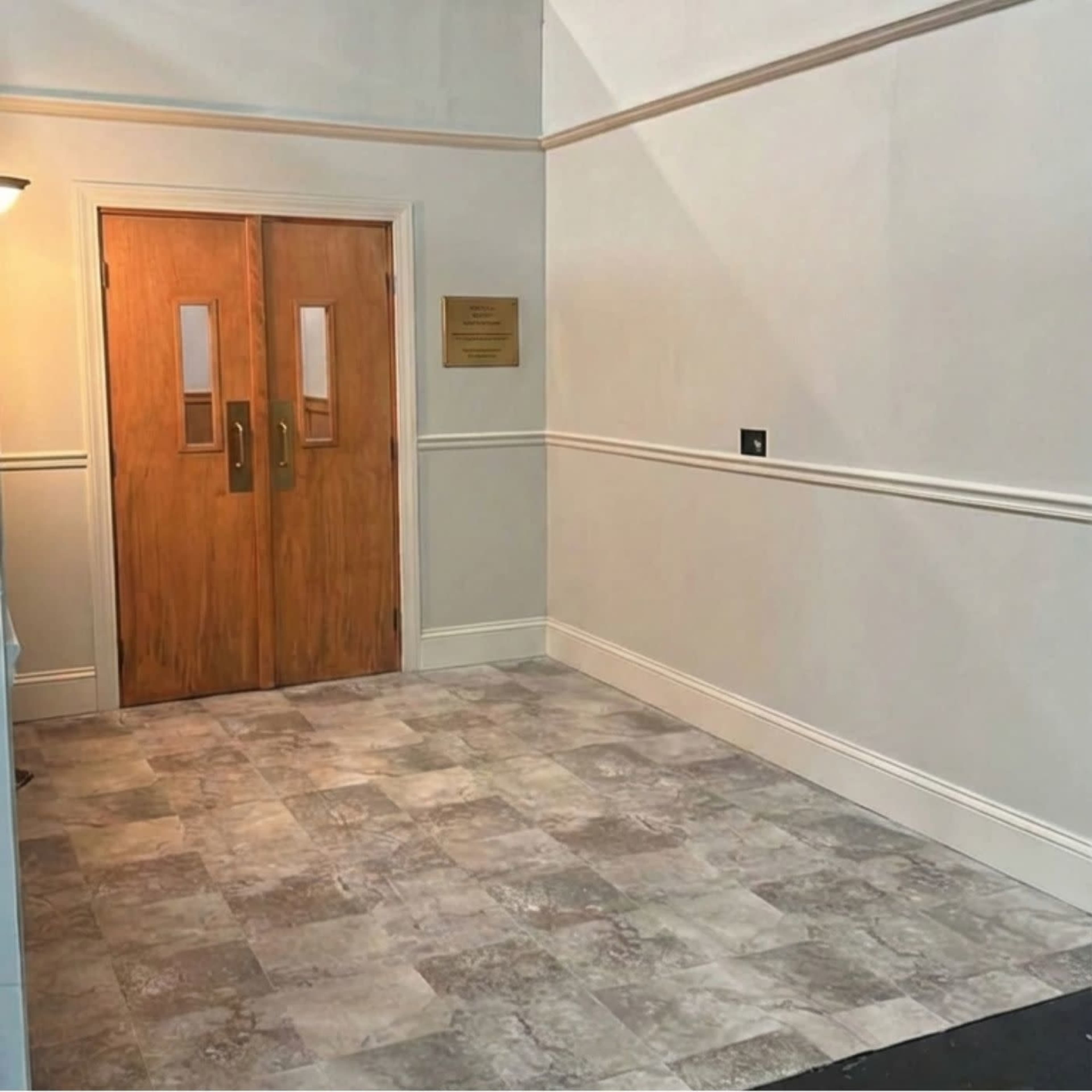 The image shows a well-lit corridor leading to a pair of wooden double doors with glass panels, surrounded by light-colored walls and a tiled floor.