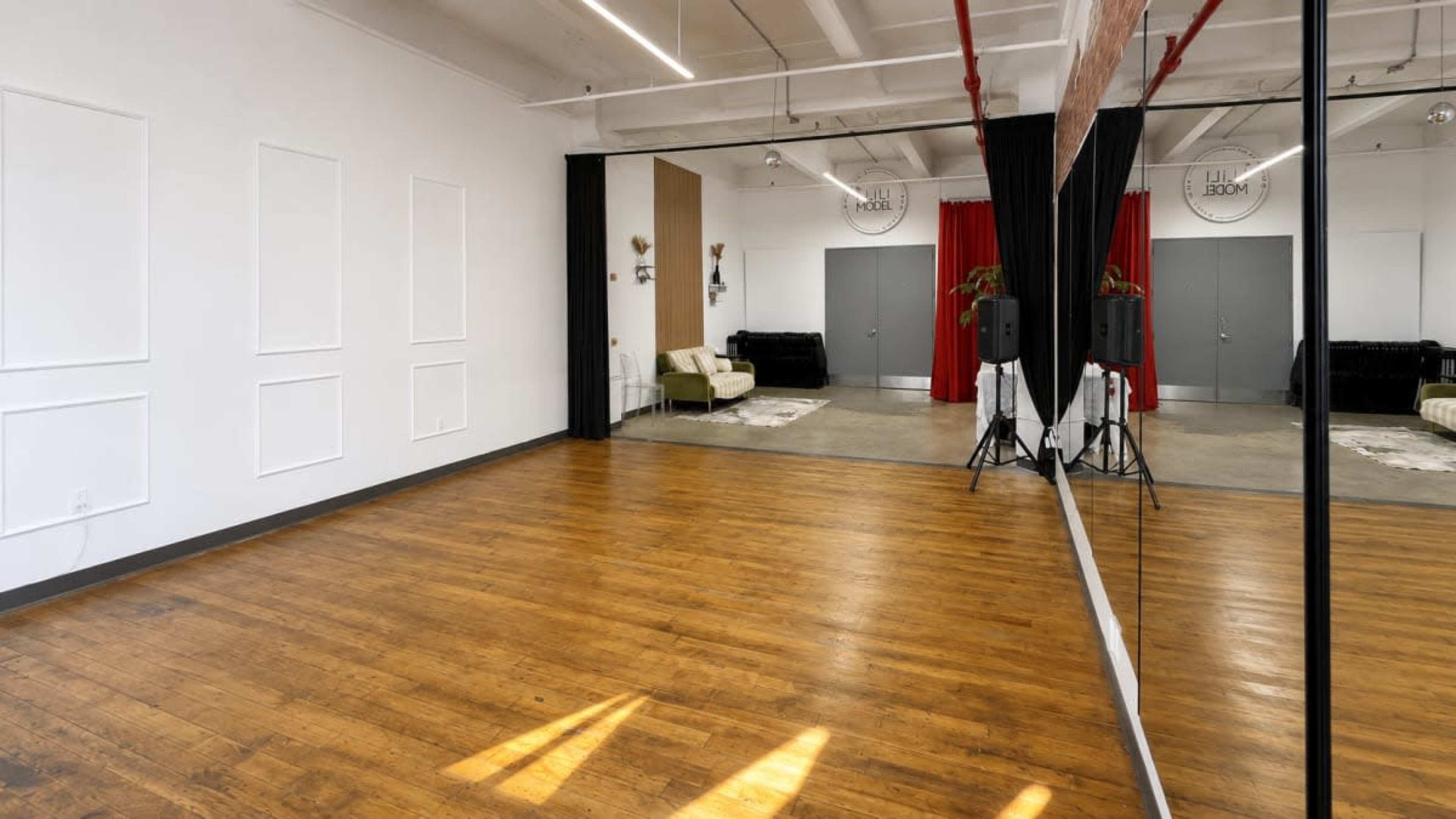 The image shows a spacious dance studio with wooden floors, large mirrors, and a black curtain separating two areas, featuring a couch and sound equipment.
