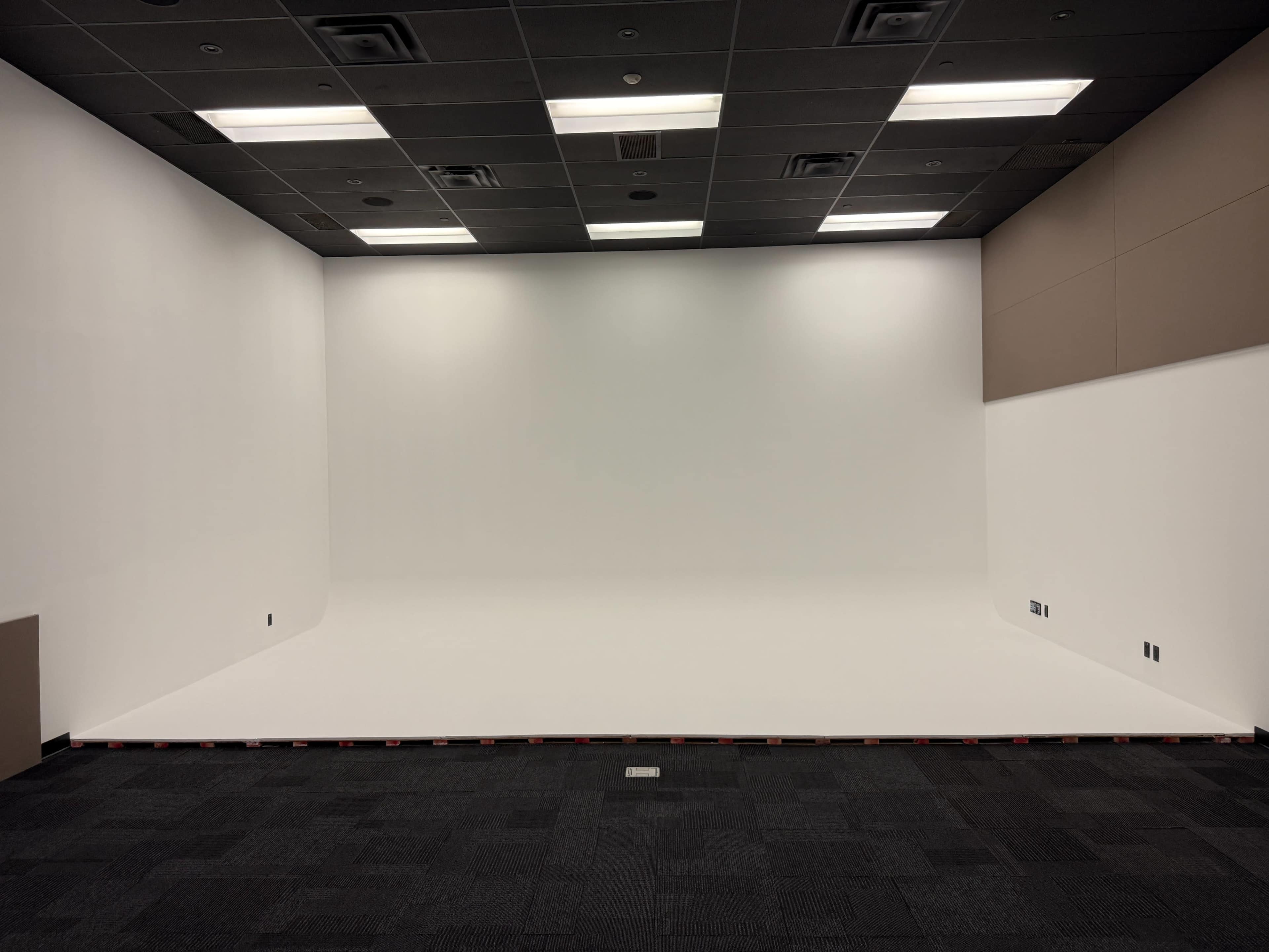 The image shows a blank, white studio space with a smooth floor and dark carpet tiles.