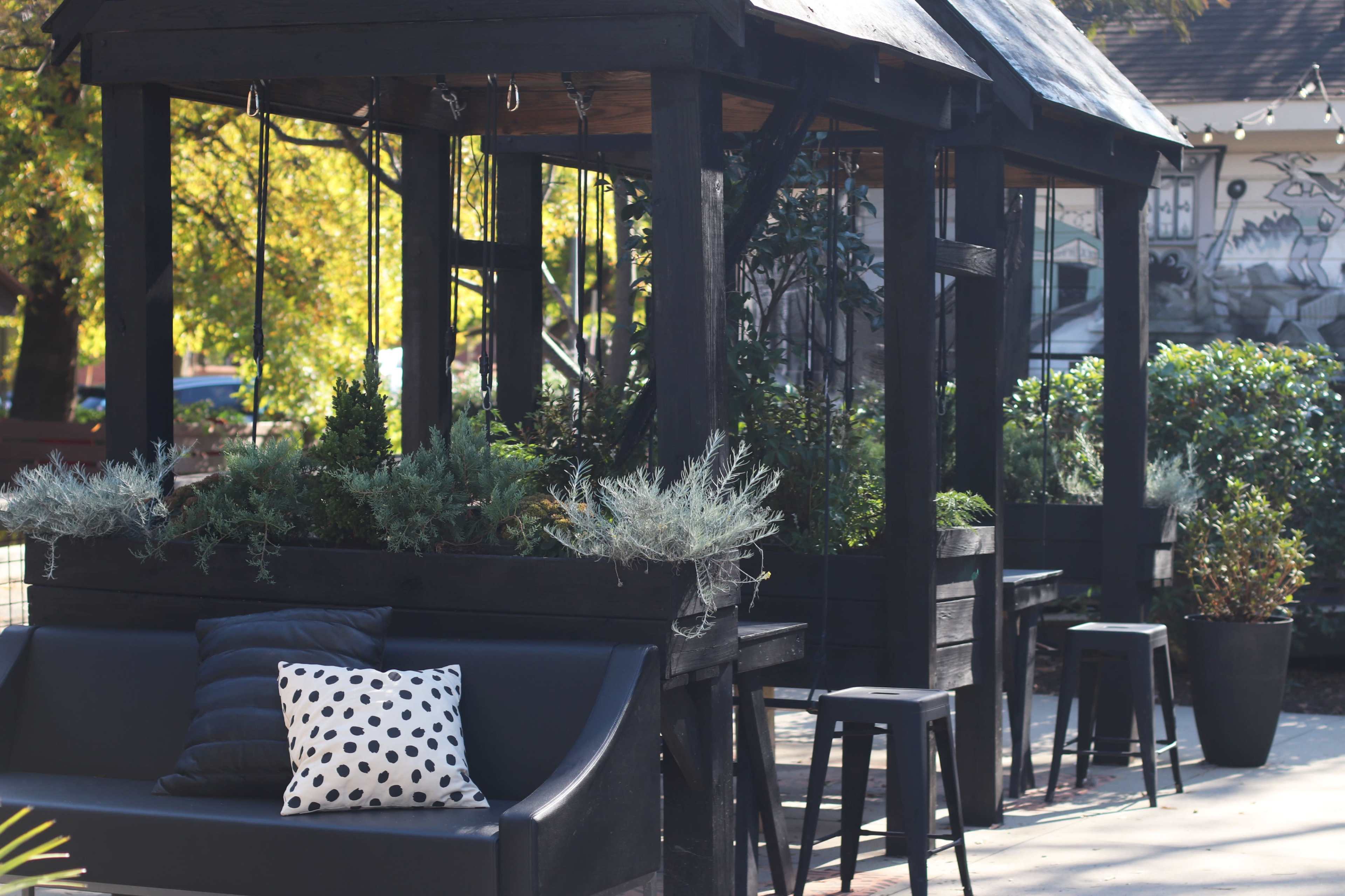 A black wooden gazebo surrounded by plants and featuring a dark sofa with a polka-dot pillow and several black stools.