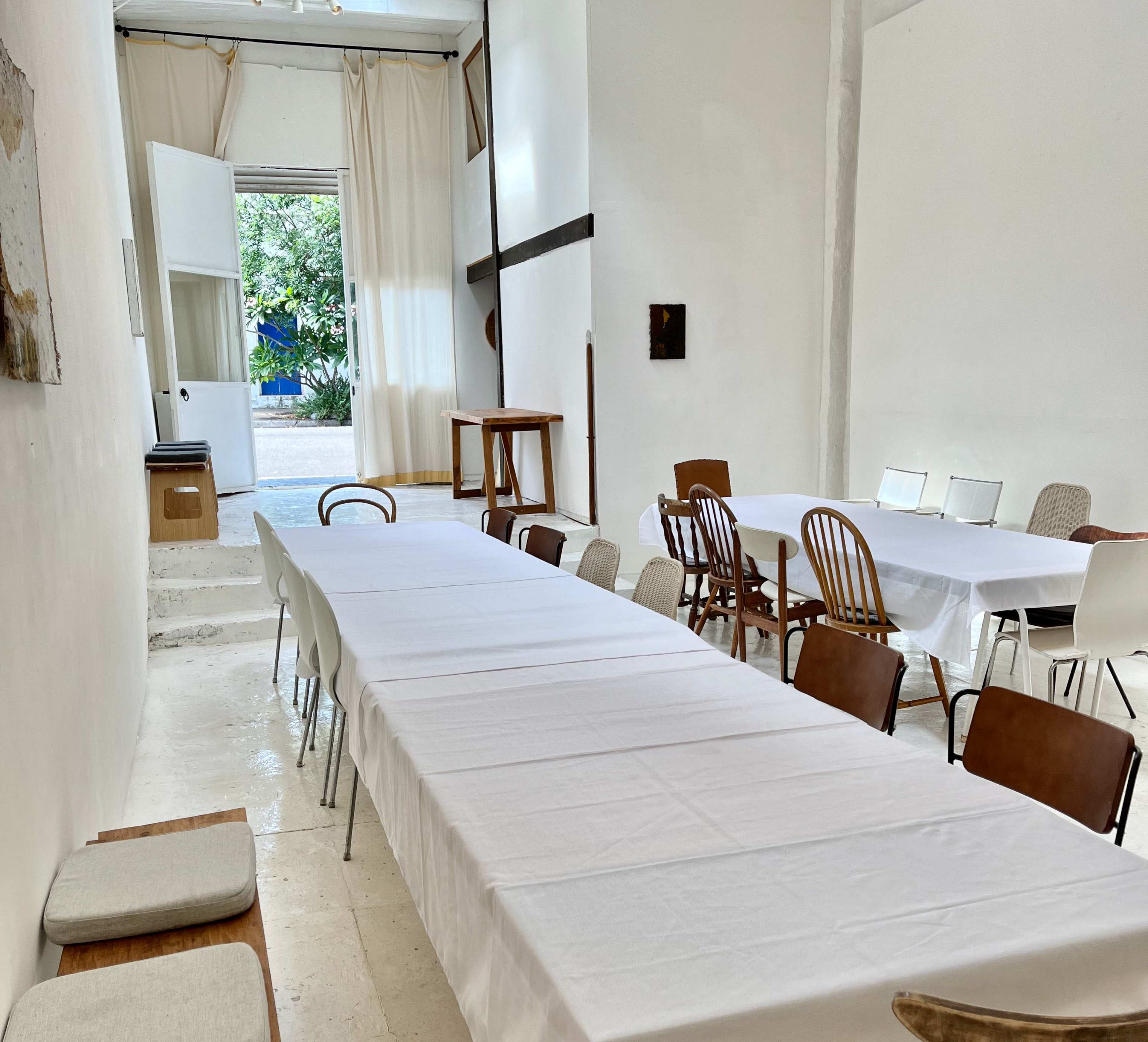 The image shows a spacious room with two long tables covered in white tablecloths, arranged with a mix of wooden and white chairs, and an open door leading to an outdoor area.