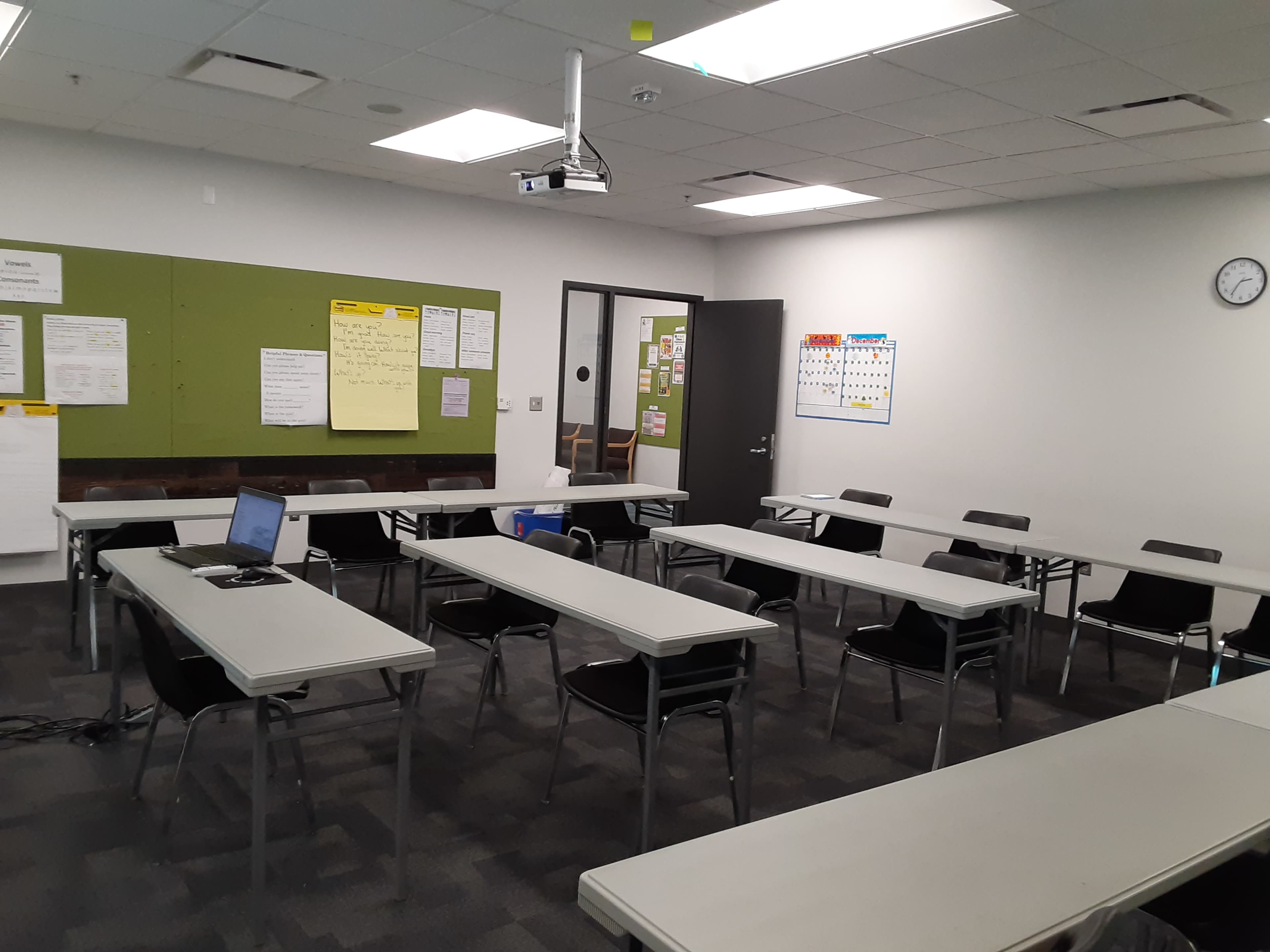 A classroom is set up with multiple tables and chairs, displaying a projector and various teaching materials on the walls.