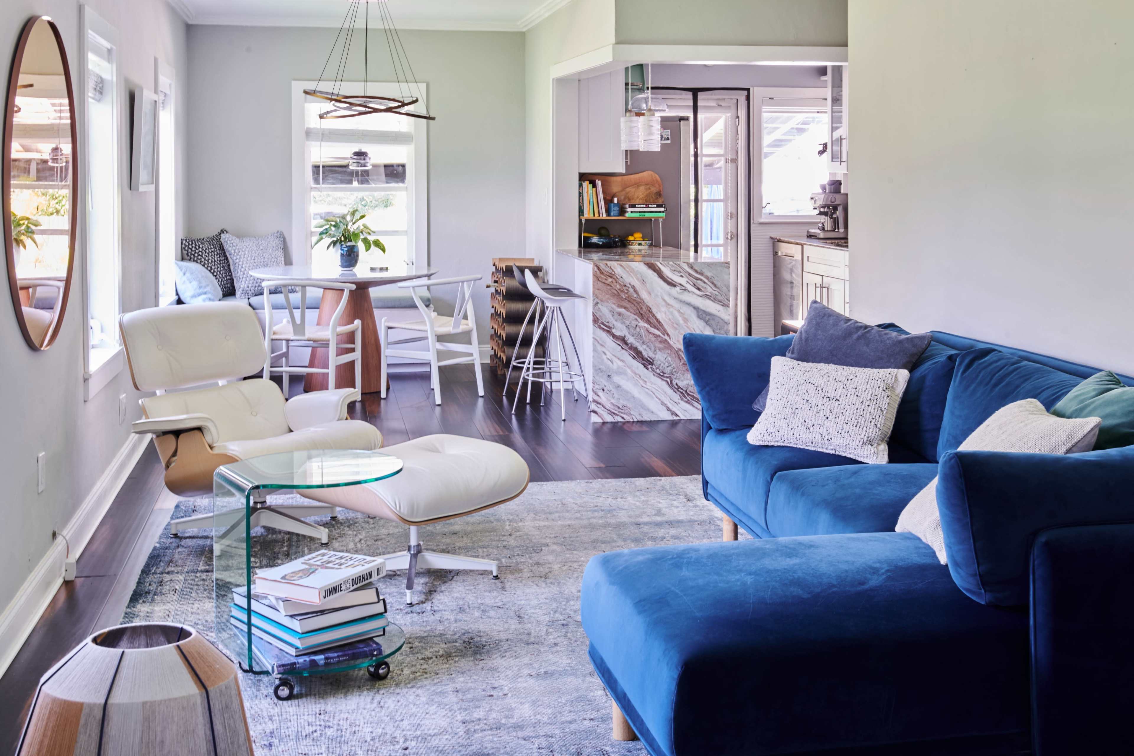 A modern living room with a blue sectional sofa, a round mirror on the wall, and a small dining area adjacent to a kitchen with marble accents.