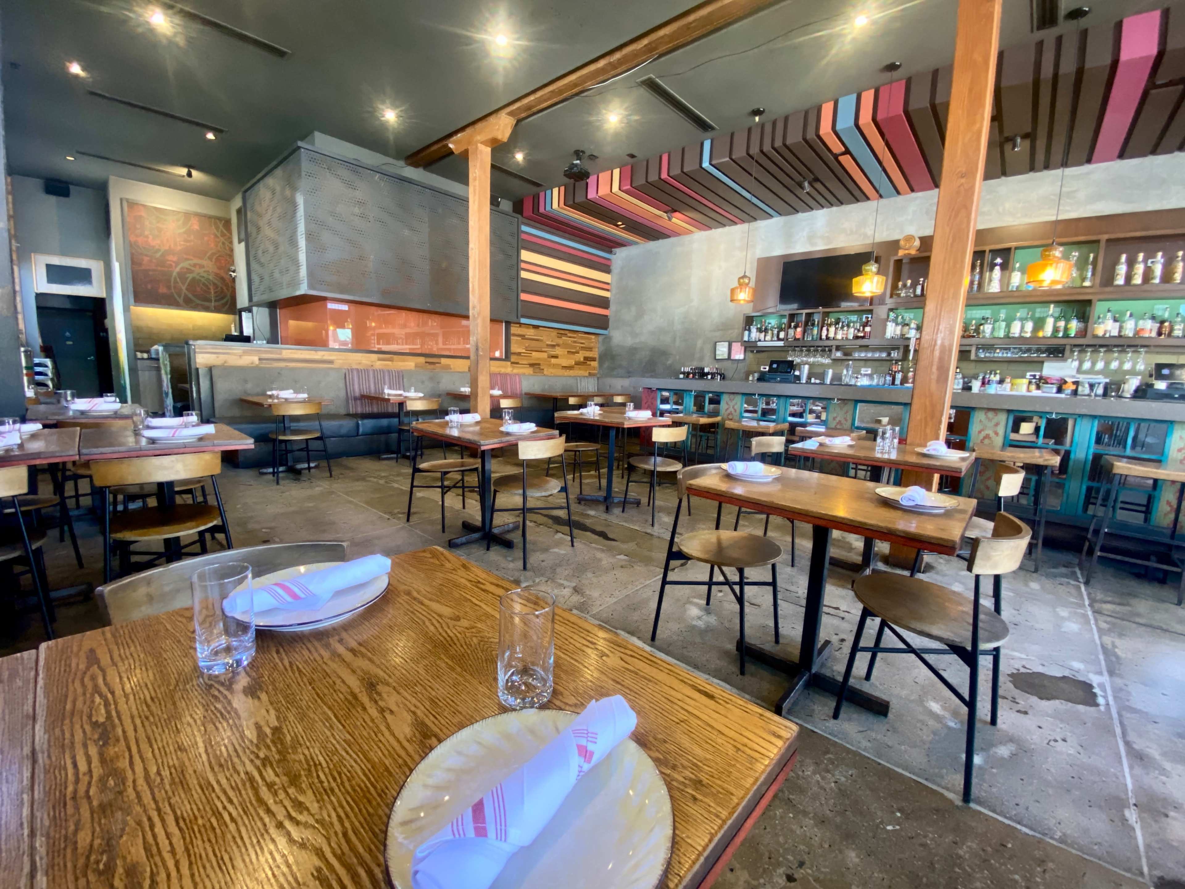 An empty restaurant interior with wooden tables, colorful wall art, and a well-stocked bar in the background.