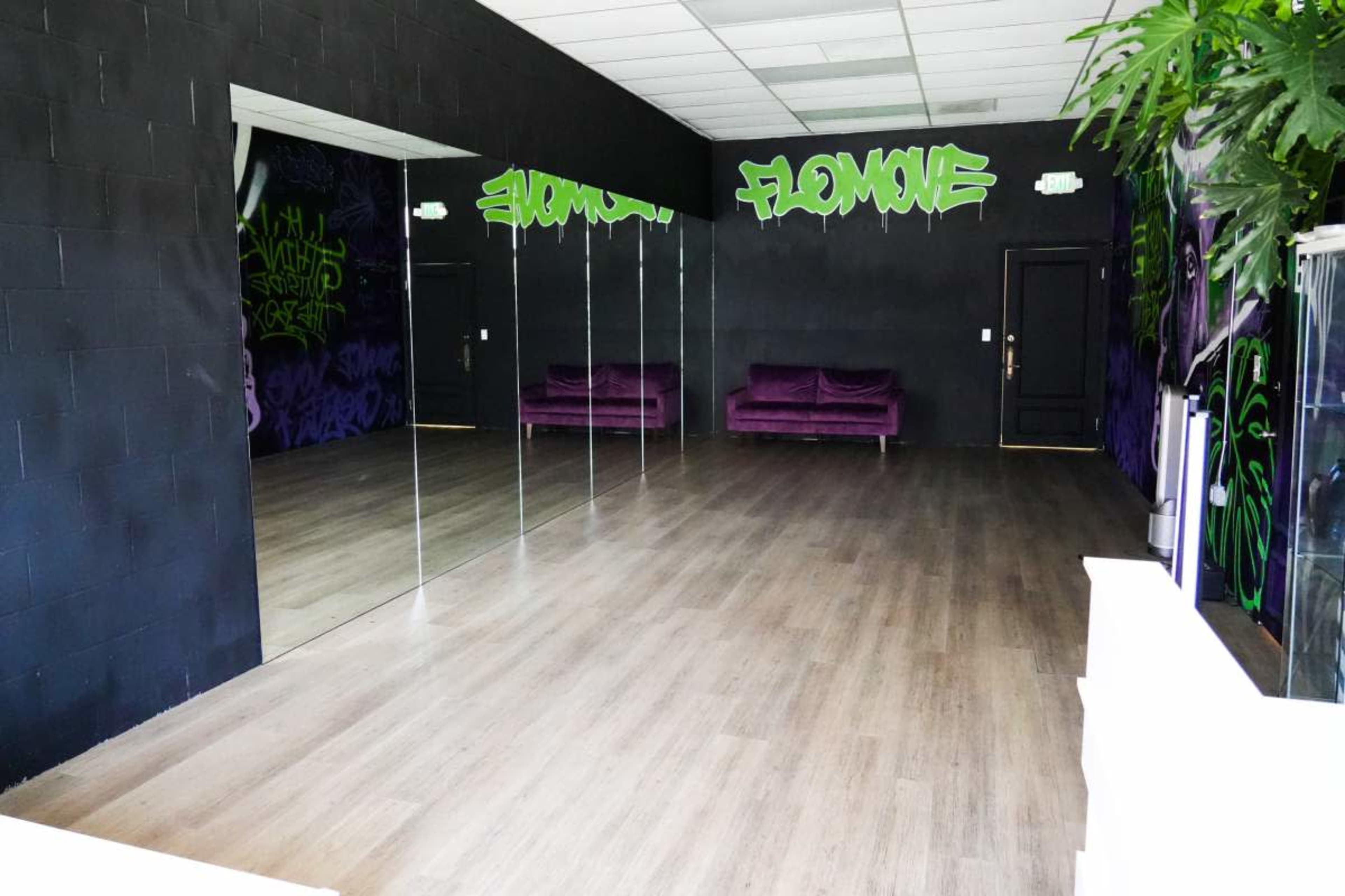 The image shows a spacious, empty room with a wooden floor, mirrored wall, and purple sofas against a black wall adorned with vibrant graffiti.