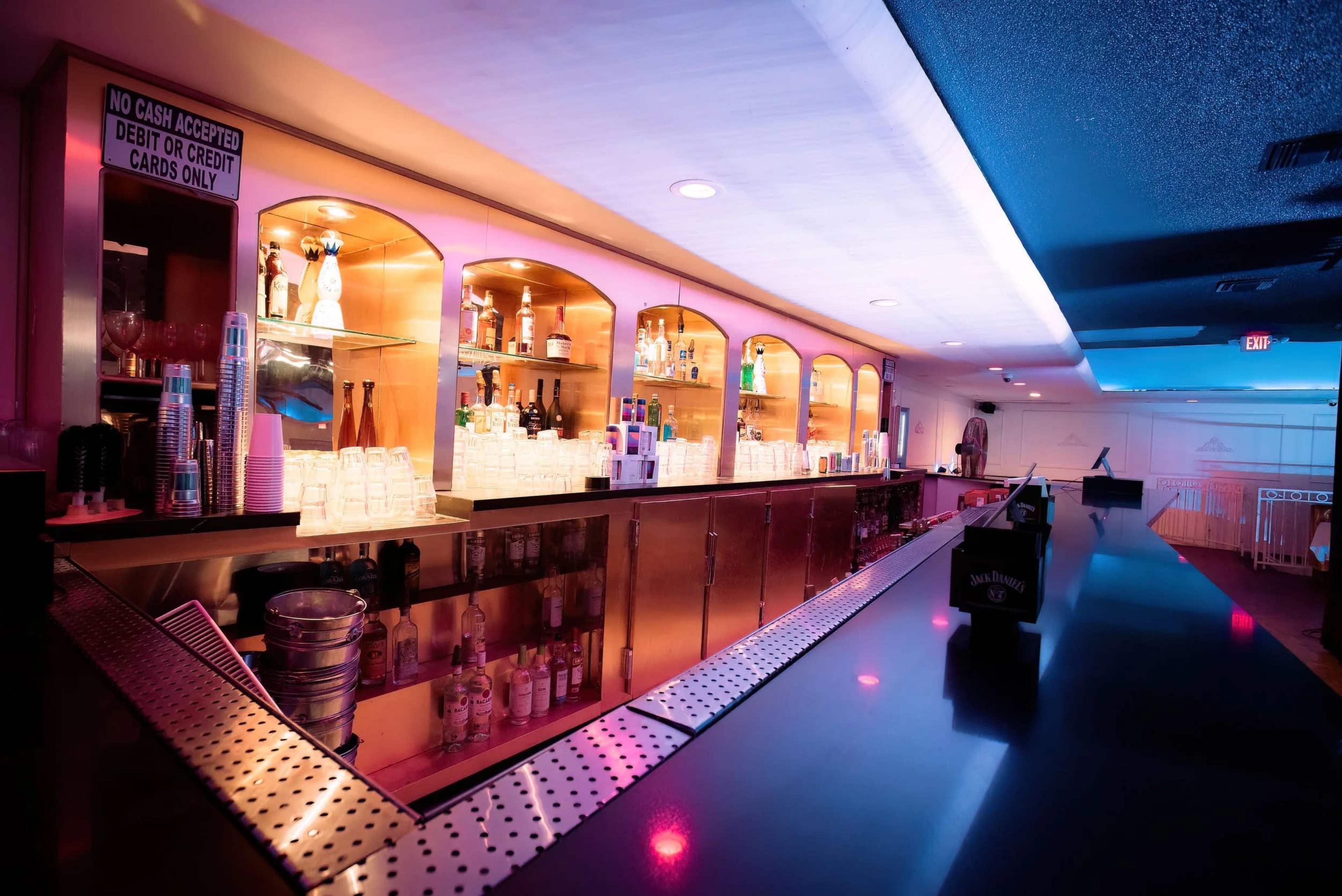 The image shows a well-lit bar with shelves displaying various bottles of alcohol and a polished countertop.