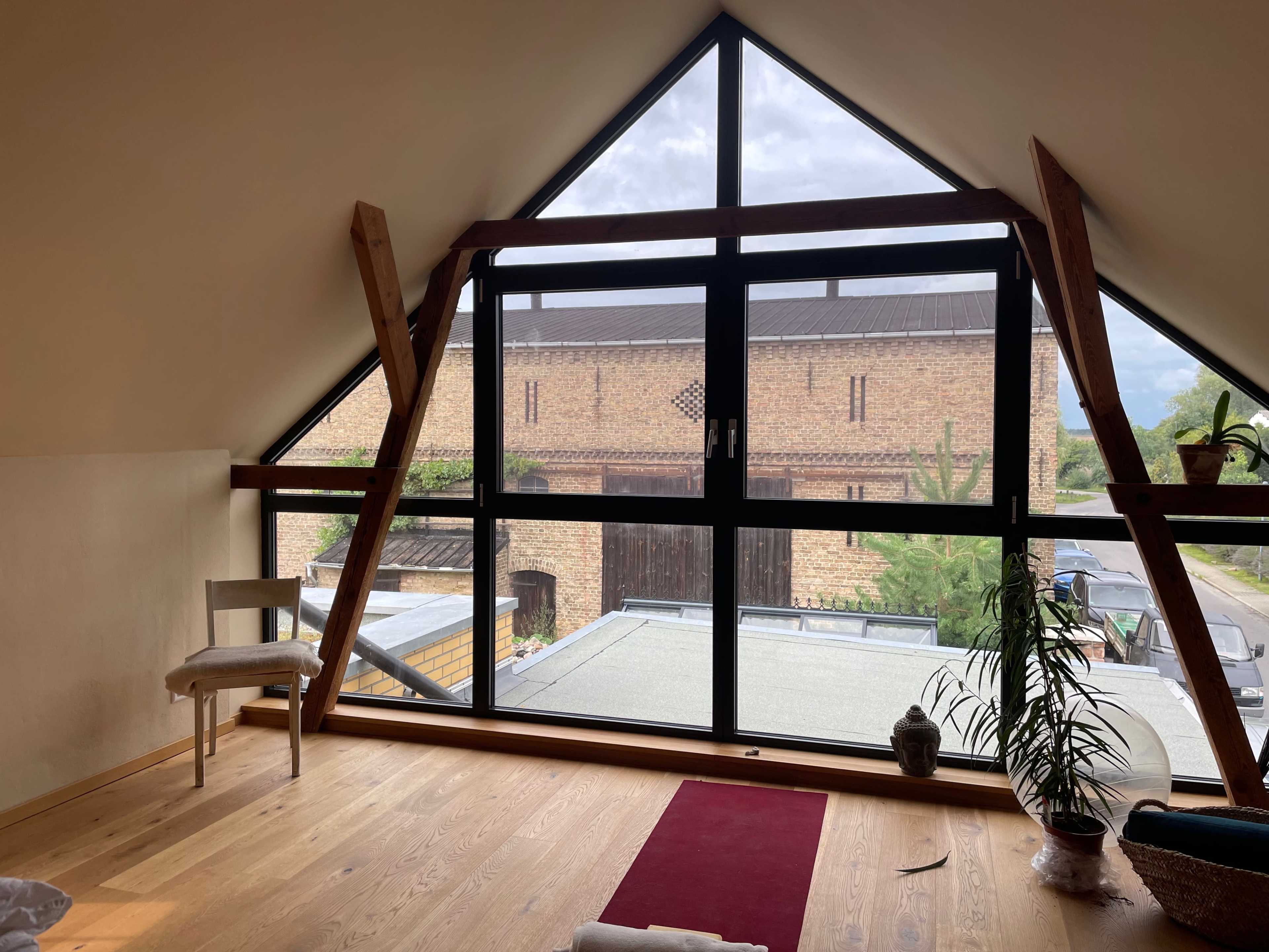 The image shows a room with a large angled window framed by wooden beams, overlooking a rustic building and a paved area outside.