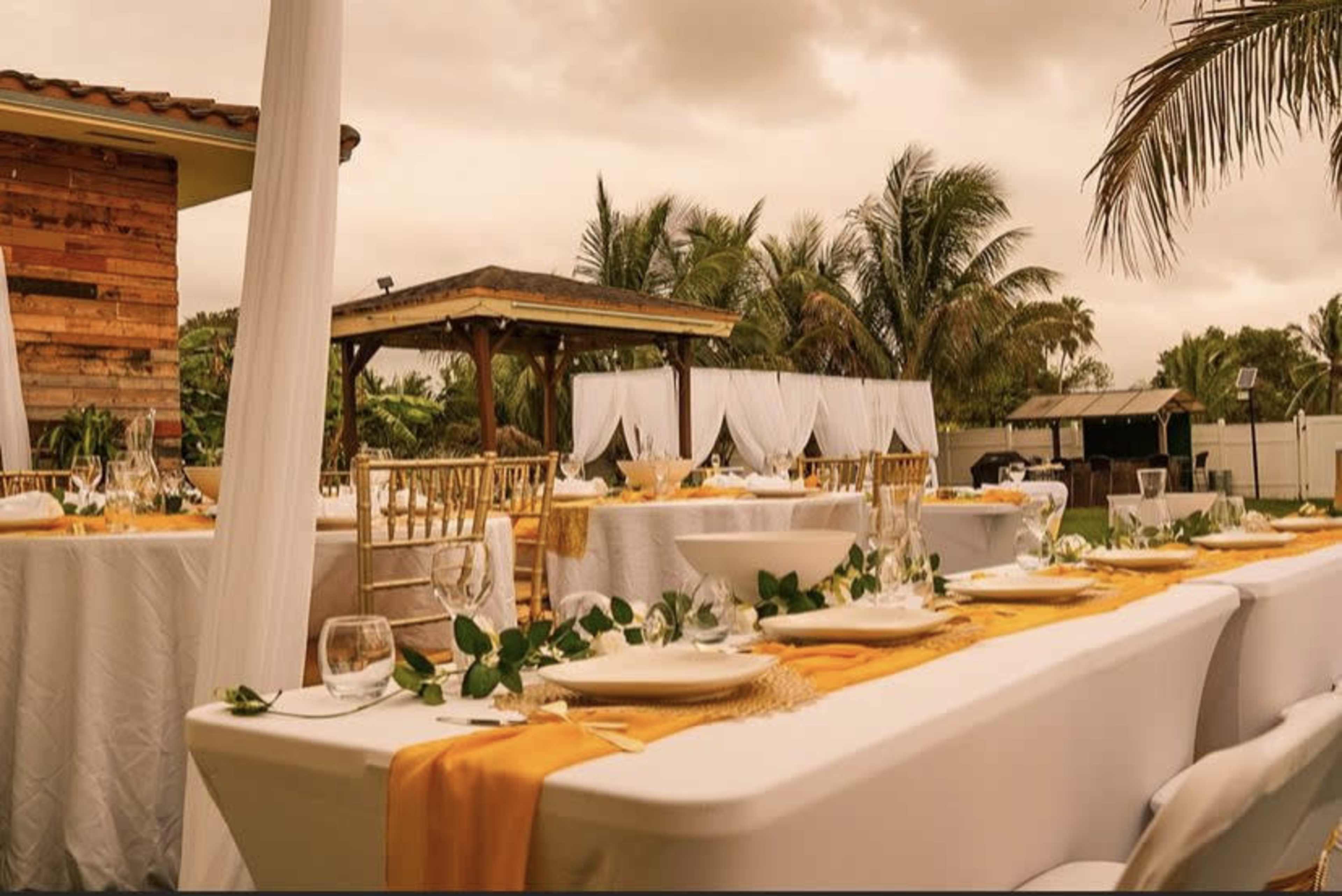 A spacious outdoor dining setup features elegantly arranged tables with white tablecloths, golden accents, and greenery, surrounded by palm trees and a gazebo.