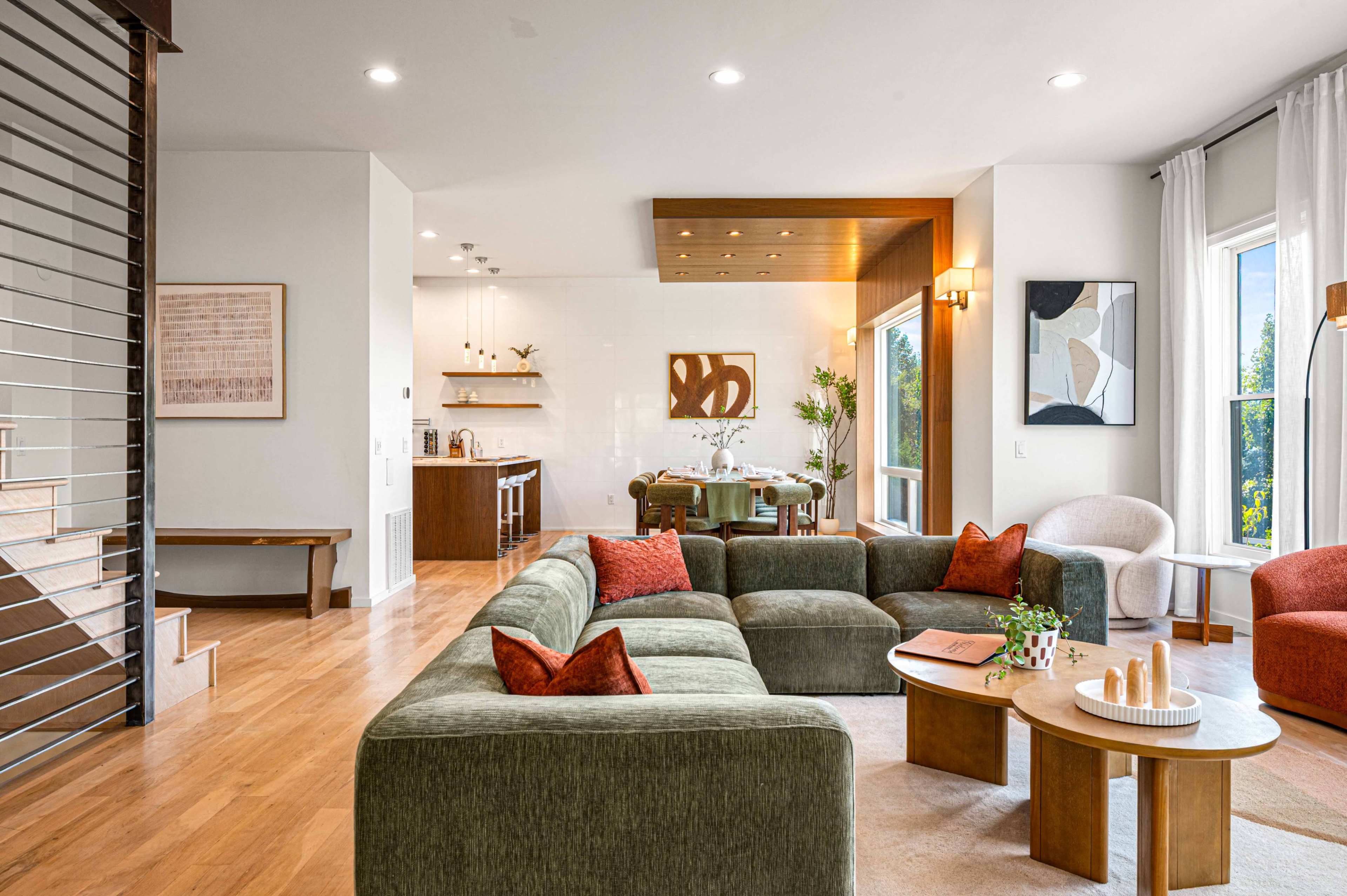 The image shows a modern living room with a spacious layout, featuring a green sectional sofa, wooden furniture, large windows, and a dining area in the background.
