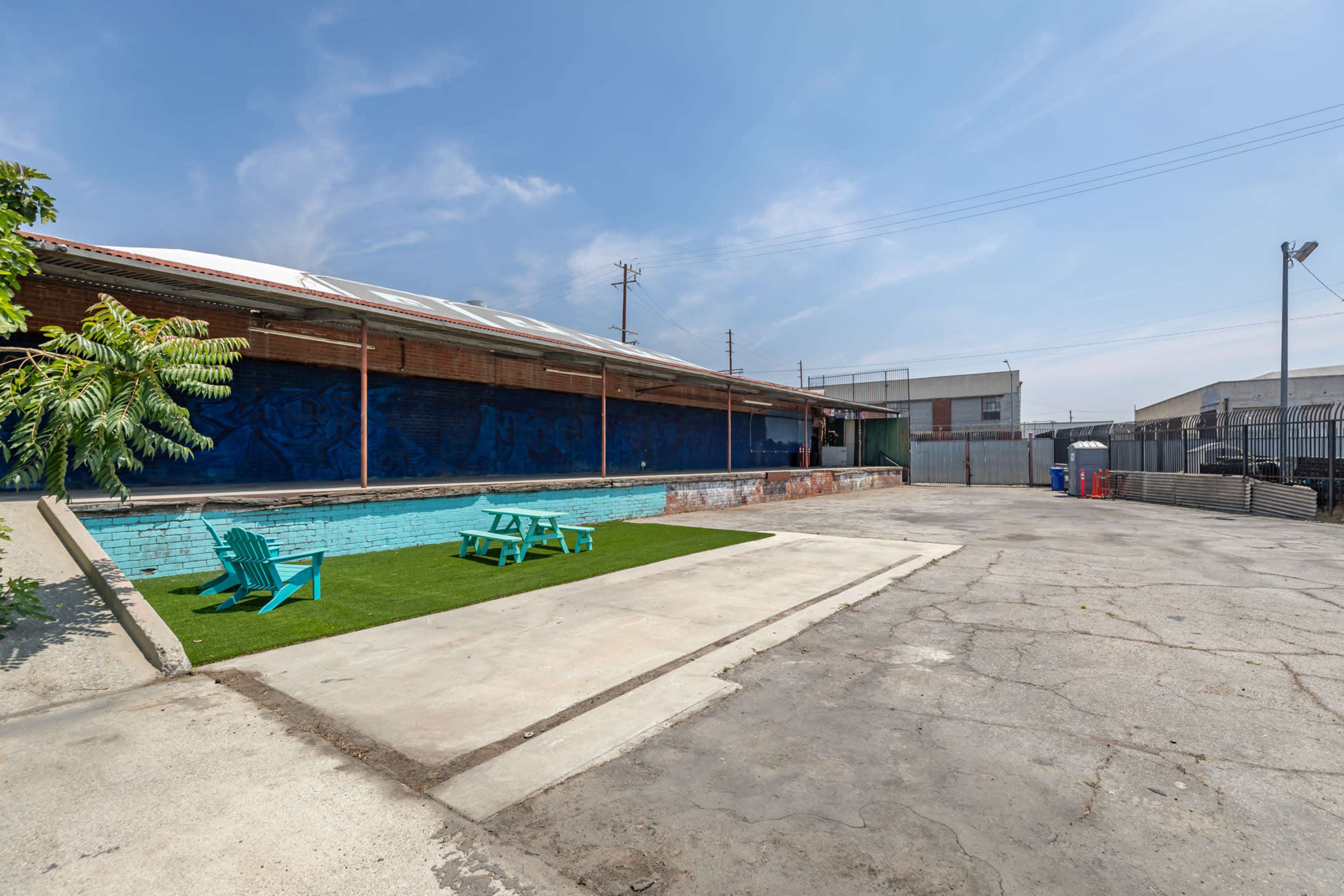 The image shows a concrete outdoor space with a small patch of artificial grass and blue adirondack chairs, adjacent to a building with a mural on its wall.