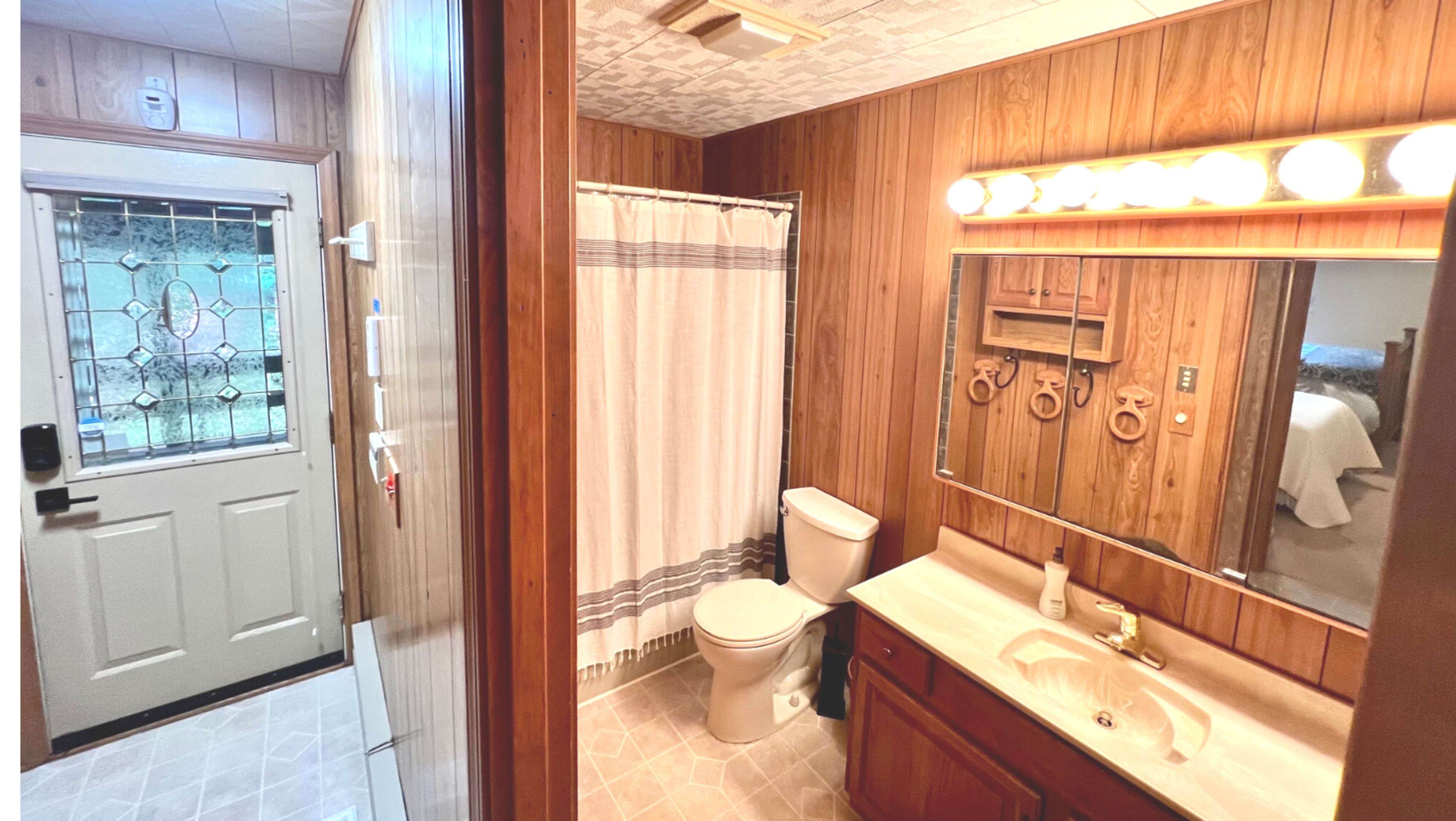 A bathroom featuring wooden paneling, a white shower curtain, a toilet, a sink with a mirror above it, and a door leading to another room.