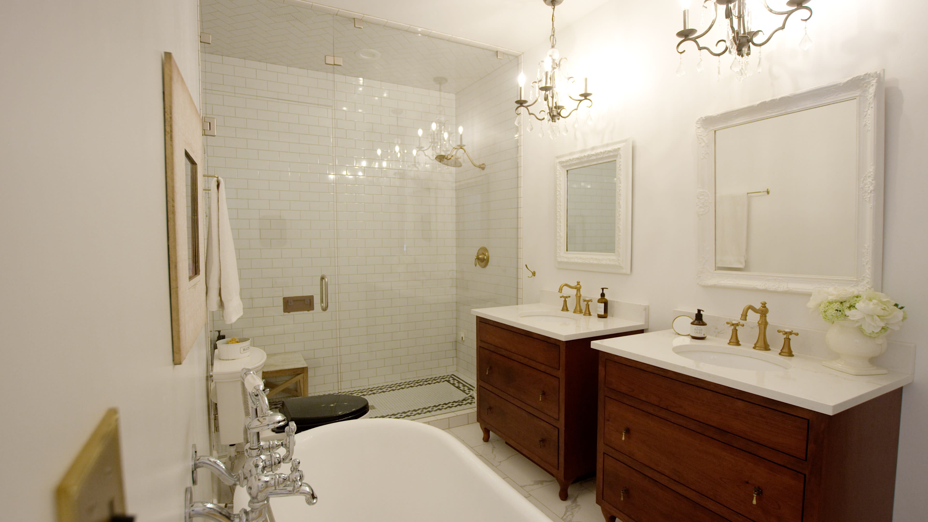 The bathroom features two wooden vanities with sinks, a freestanding bathtub, and a glass shower enclosed by white tiled walls.