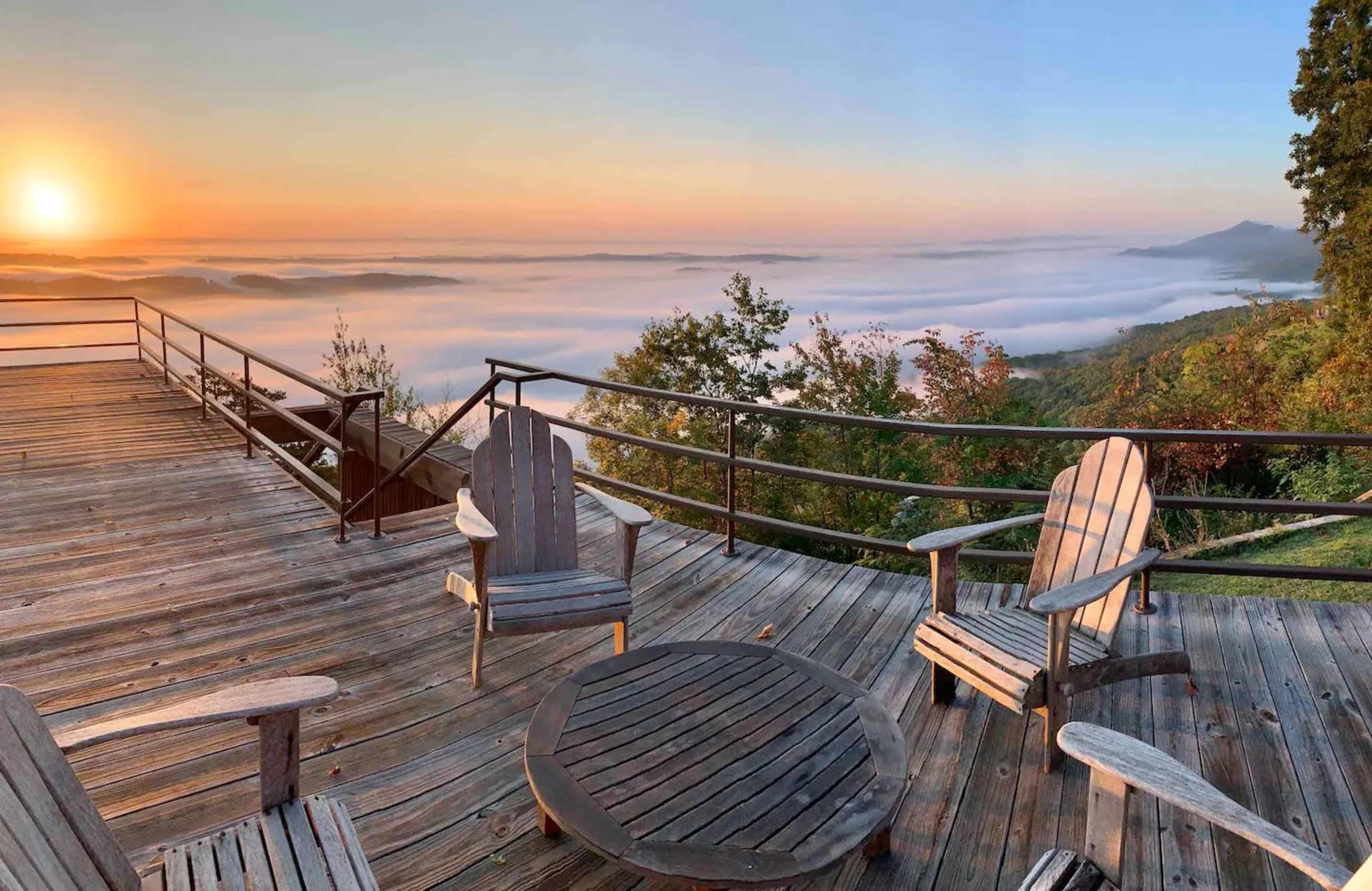 A wooden deck with four chairs and a round table overlooks a landscape shrouded in fog at sunrise.