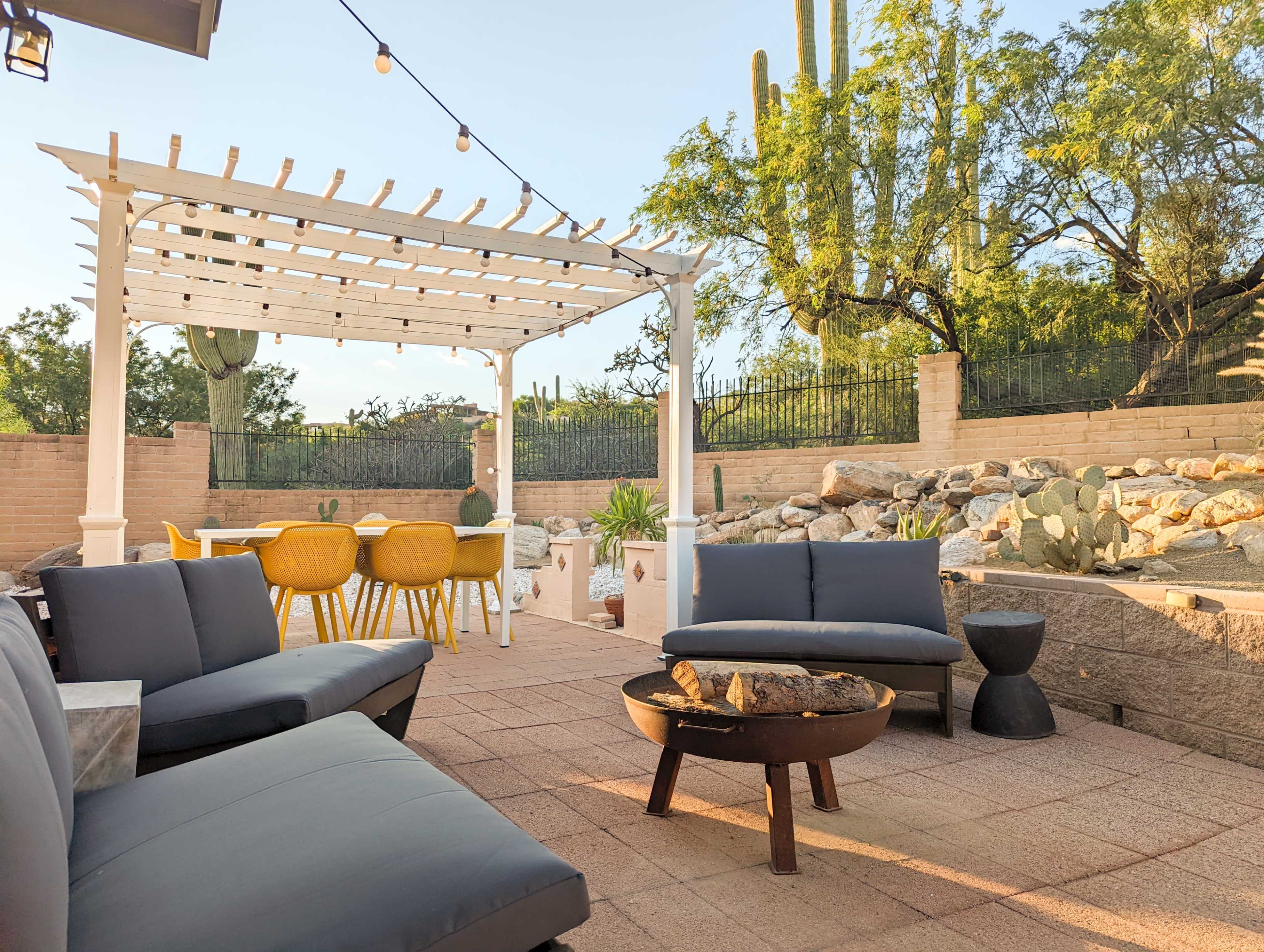 A patio area features modern seating, a fire pit, a dining table, and cacti in a landscaped yard under a clear sky.