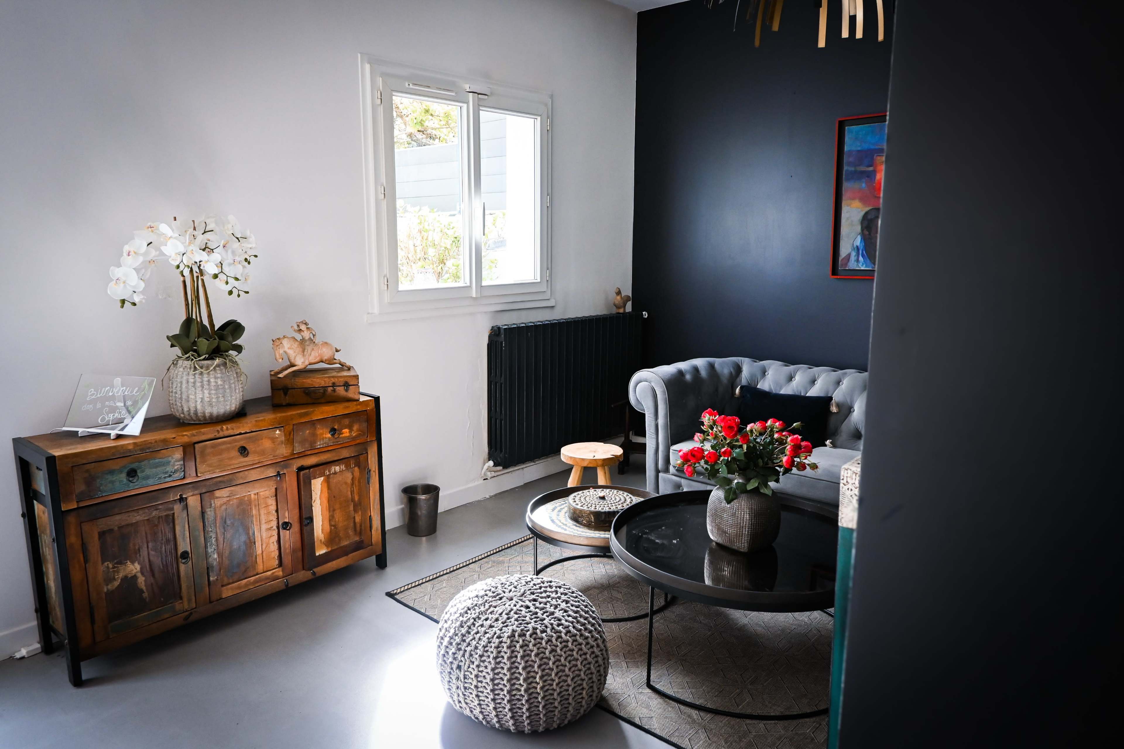 The image shows a modern living room with a gray chesterfield sofa, a wooden sideboard, a round coffee table, and decorative plants.