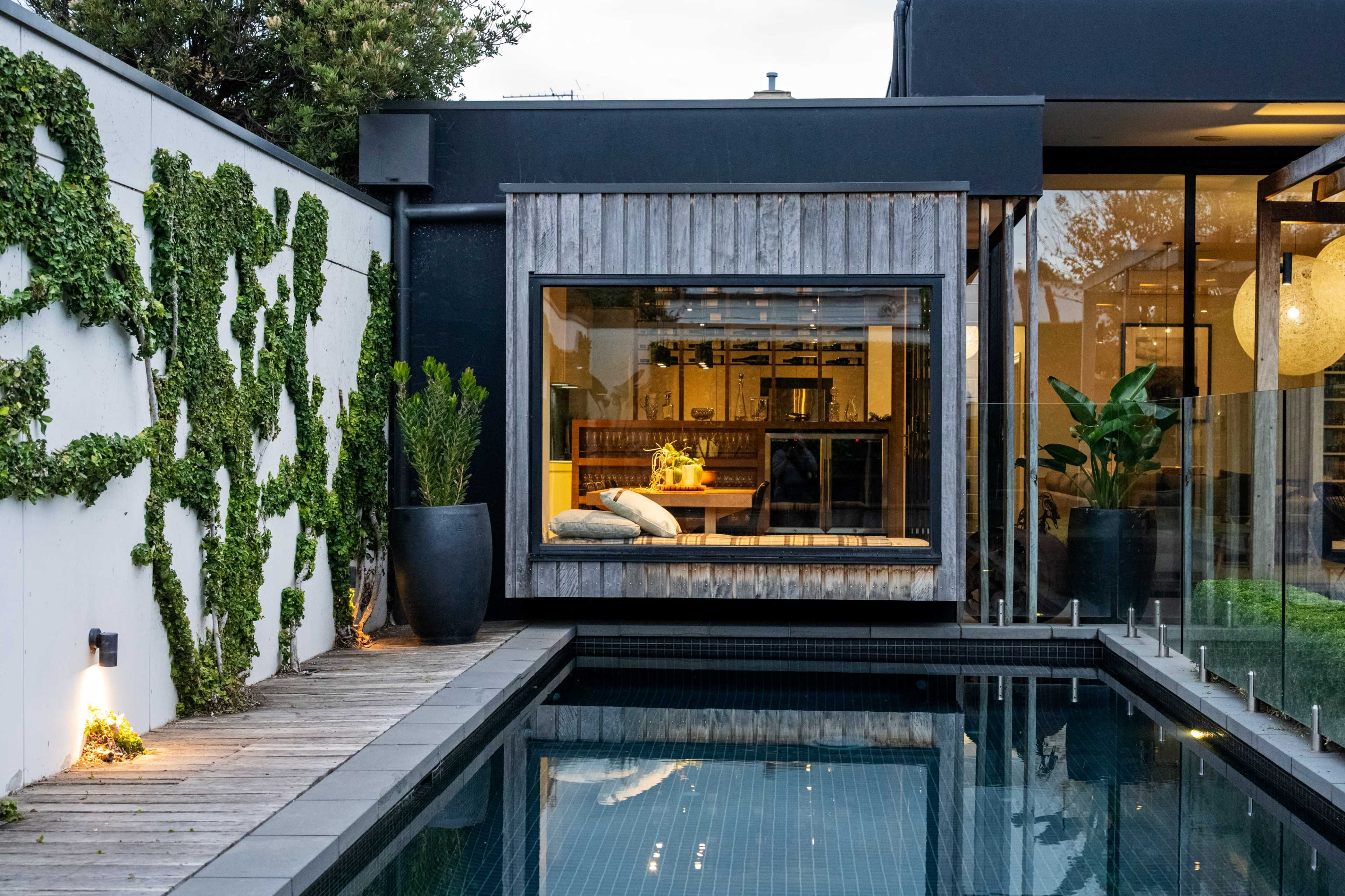 A modern home features a sleek swimming pool alongside a wooden deck and a window showcasing a warmly lit interior.