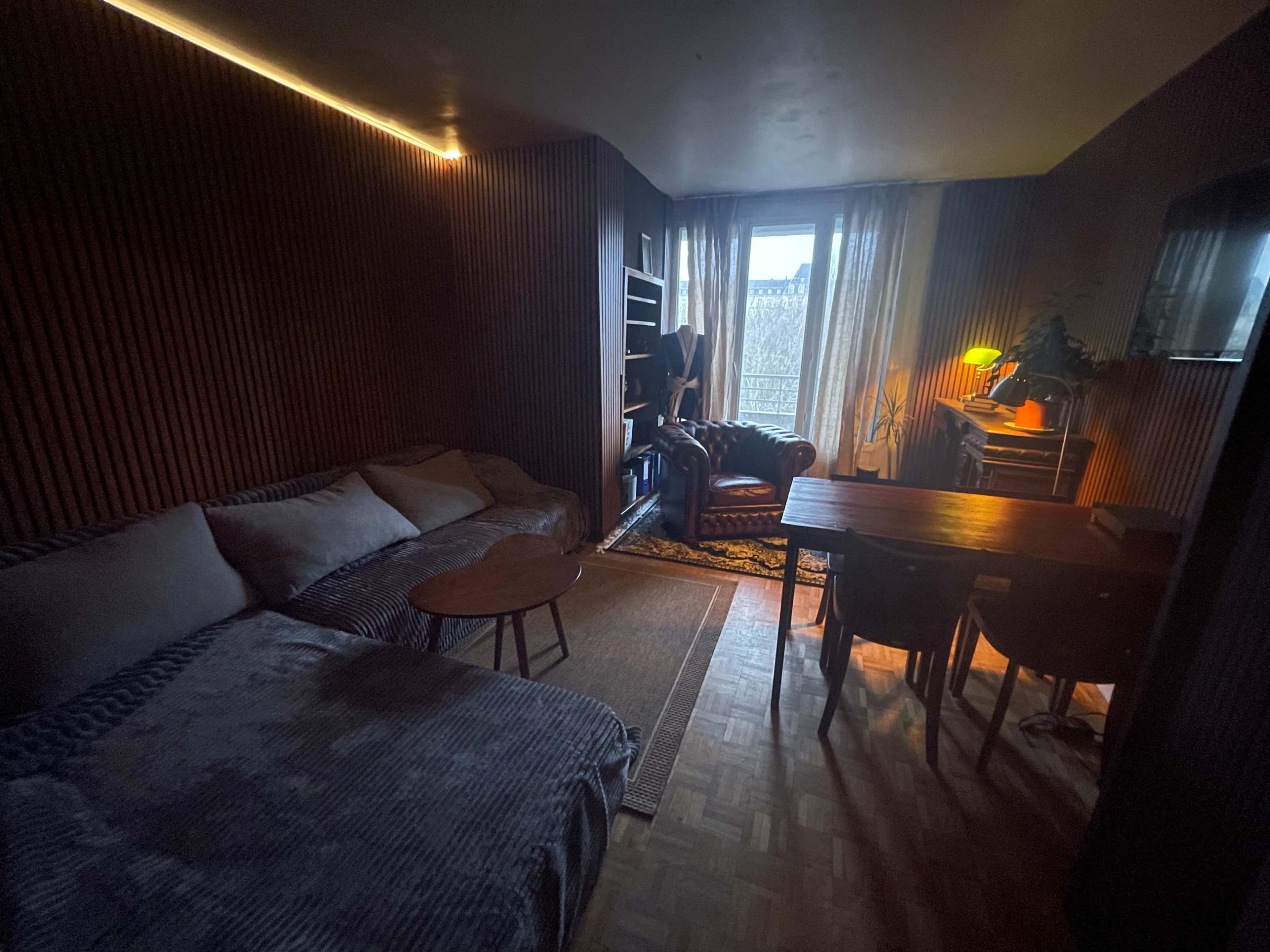 The image shows a small living area featuring a sofa, a wooden table with chairs, a bookshelf, and a window with sheer curtains.