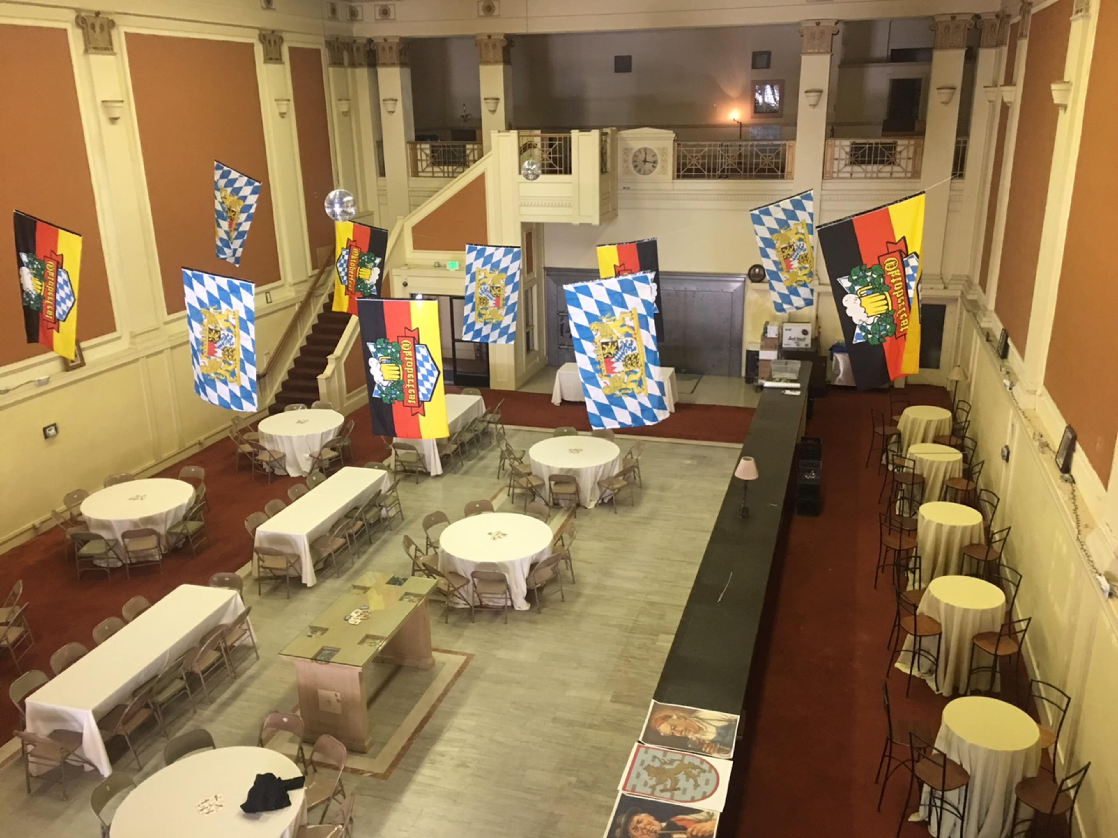 The interior of a spacious hall features tables arranged for an event, with colorful banners hanging from the ceiling and a bar area along one side.