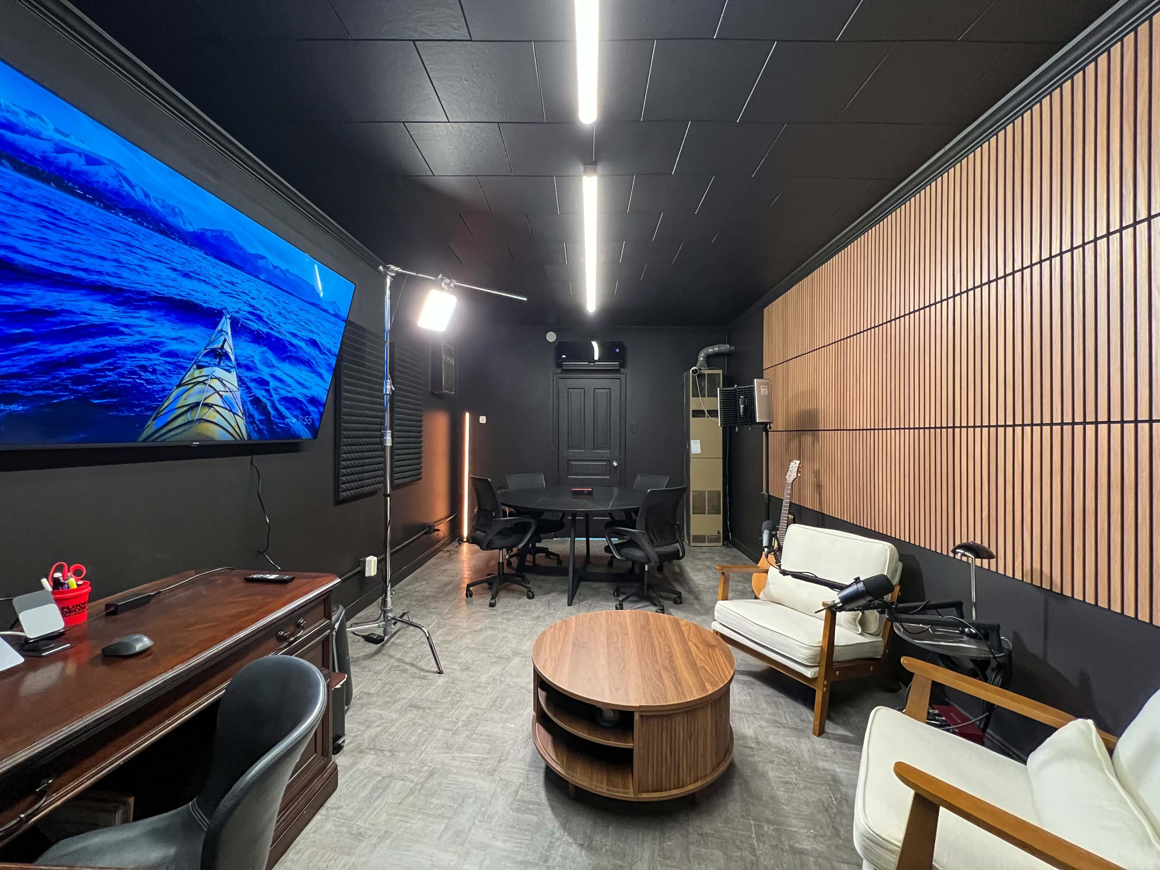 The image shows a modern interior of a small office or meeting room featuring a wooden desk, a round coffee table, seating, and a large screen displaying a kayak on water.