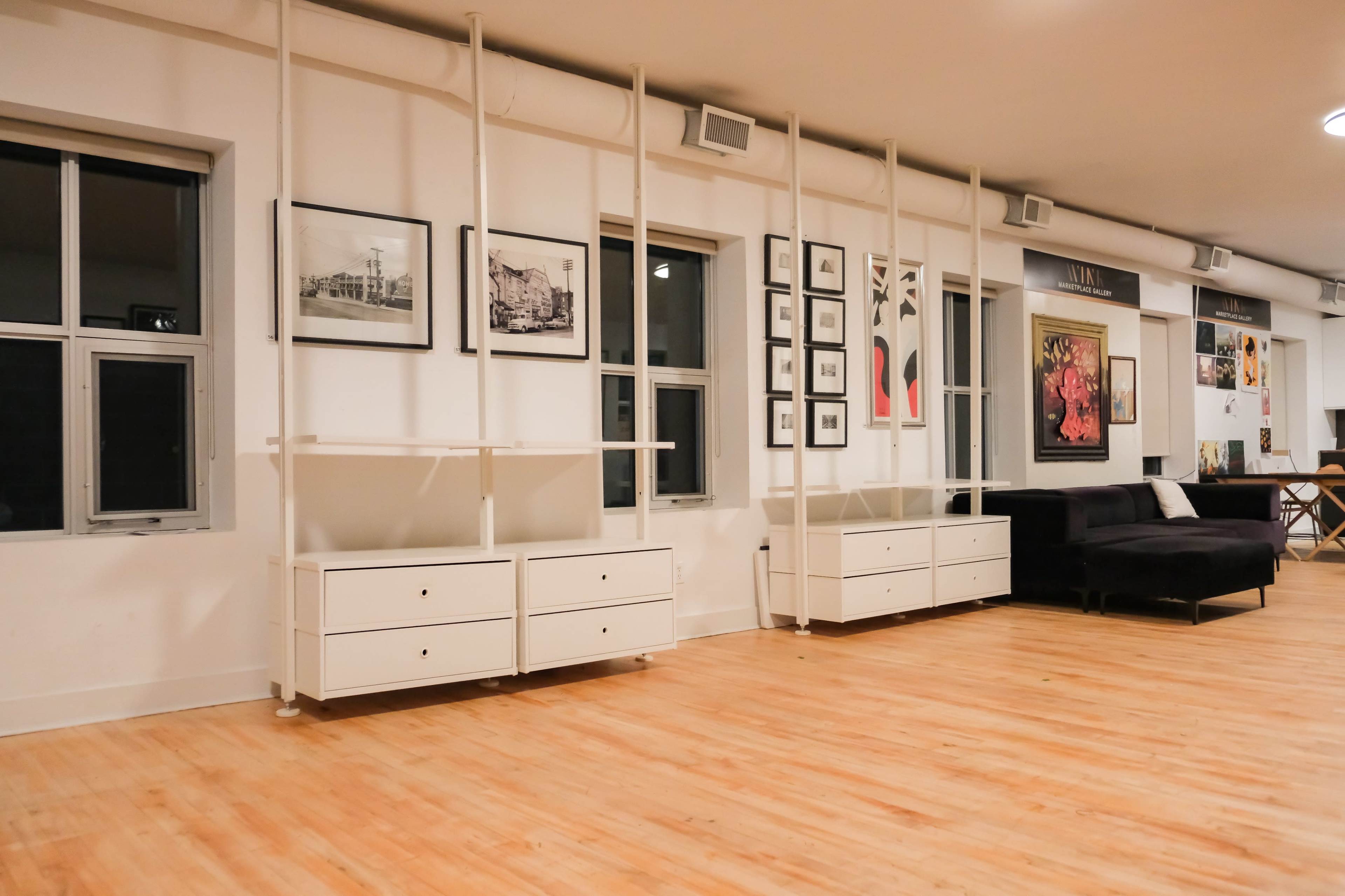 The image shows a spacious interior with white walls, wooden flooring, and shelving units against the walls, adorned with black-and-white photographs and colorful framed artwork.