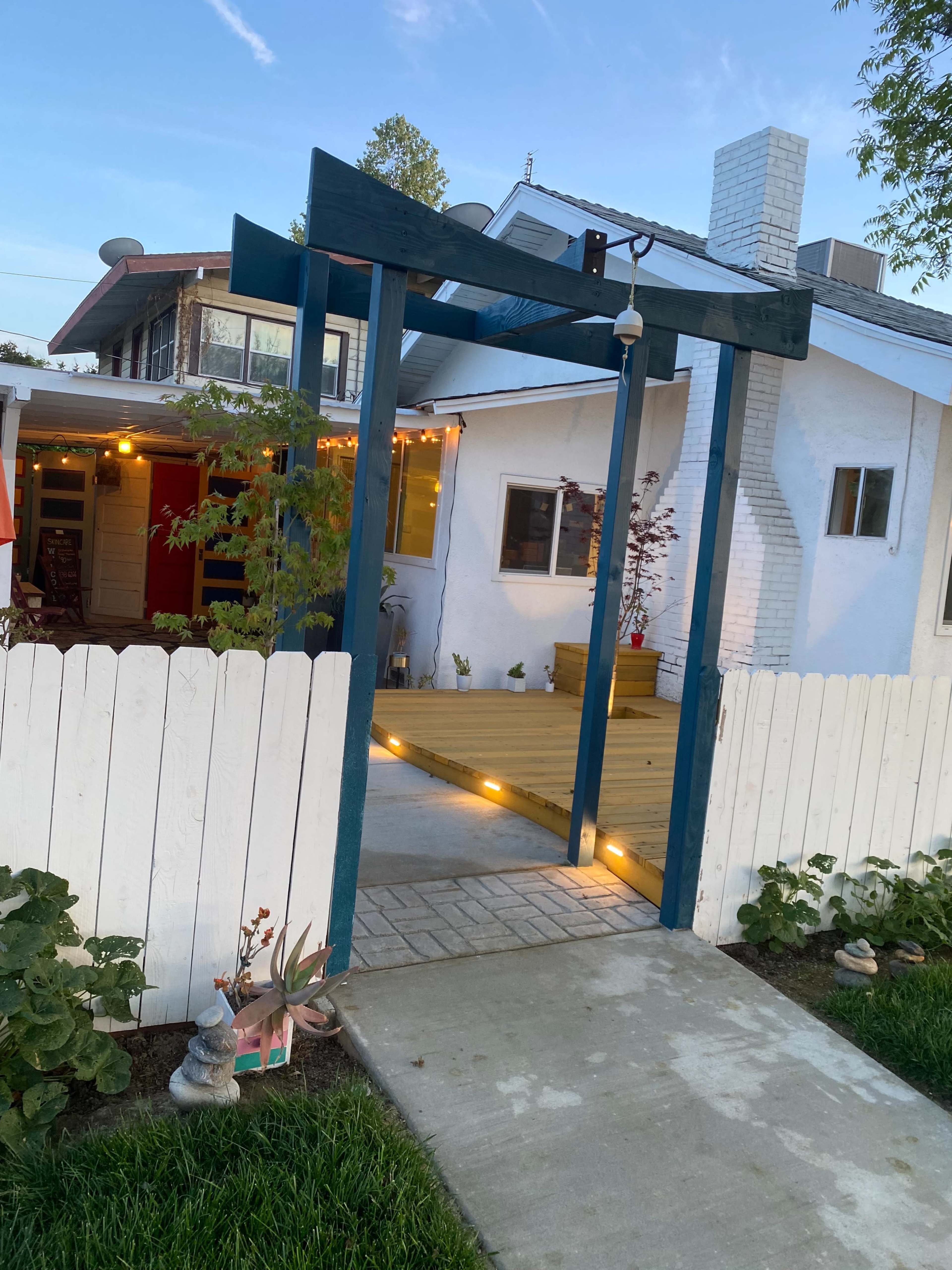 A wooden archway with a lantern leads to a pathway and deck area beside a white house, enclosed by a white picket fence.