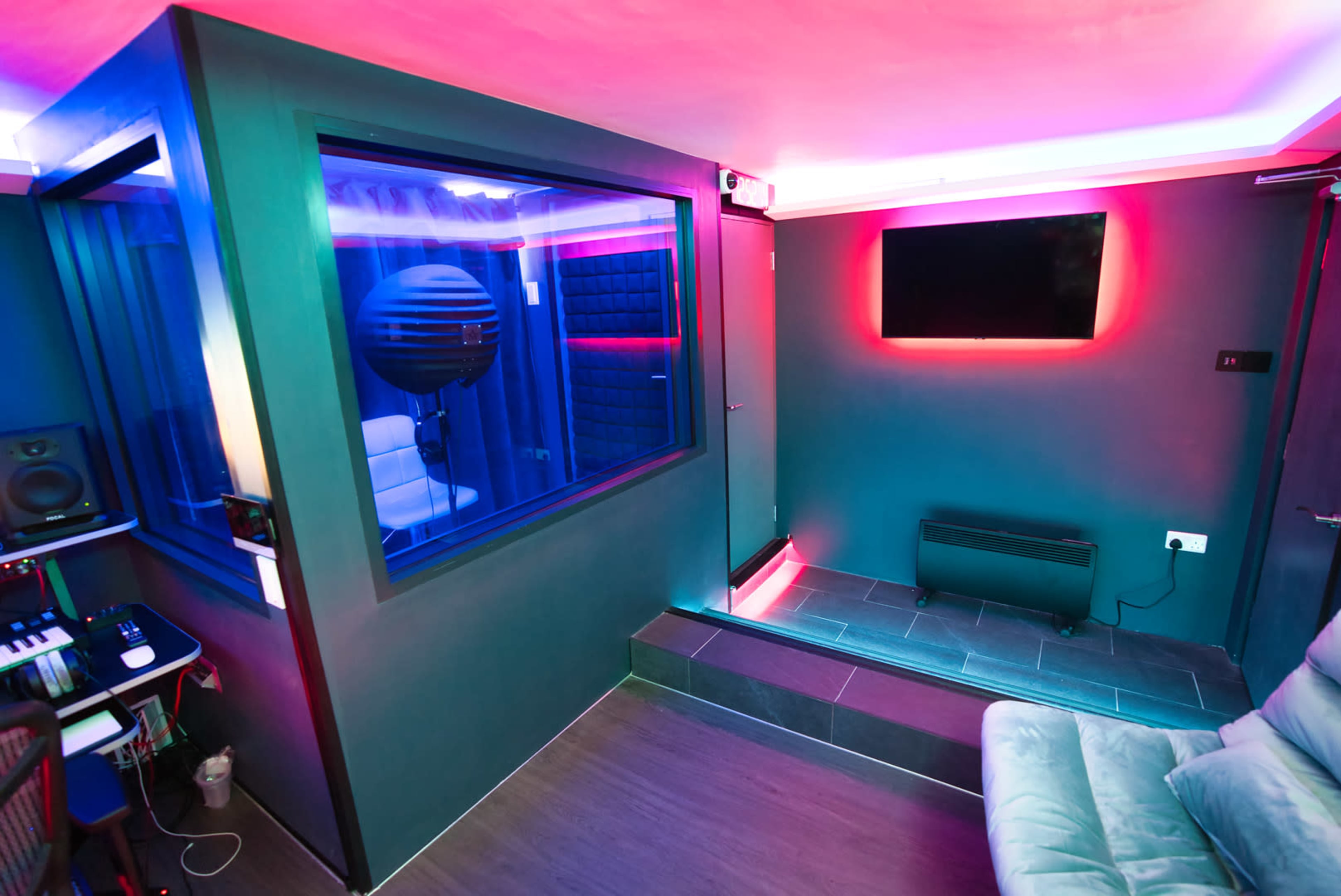 A modern music studio featuring a soundproof booth, a television mounted on the wall, and a cozy seating area illuminated by colored LED lights.