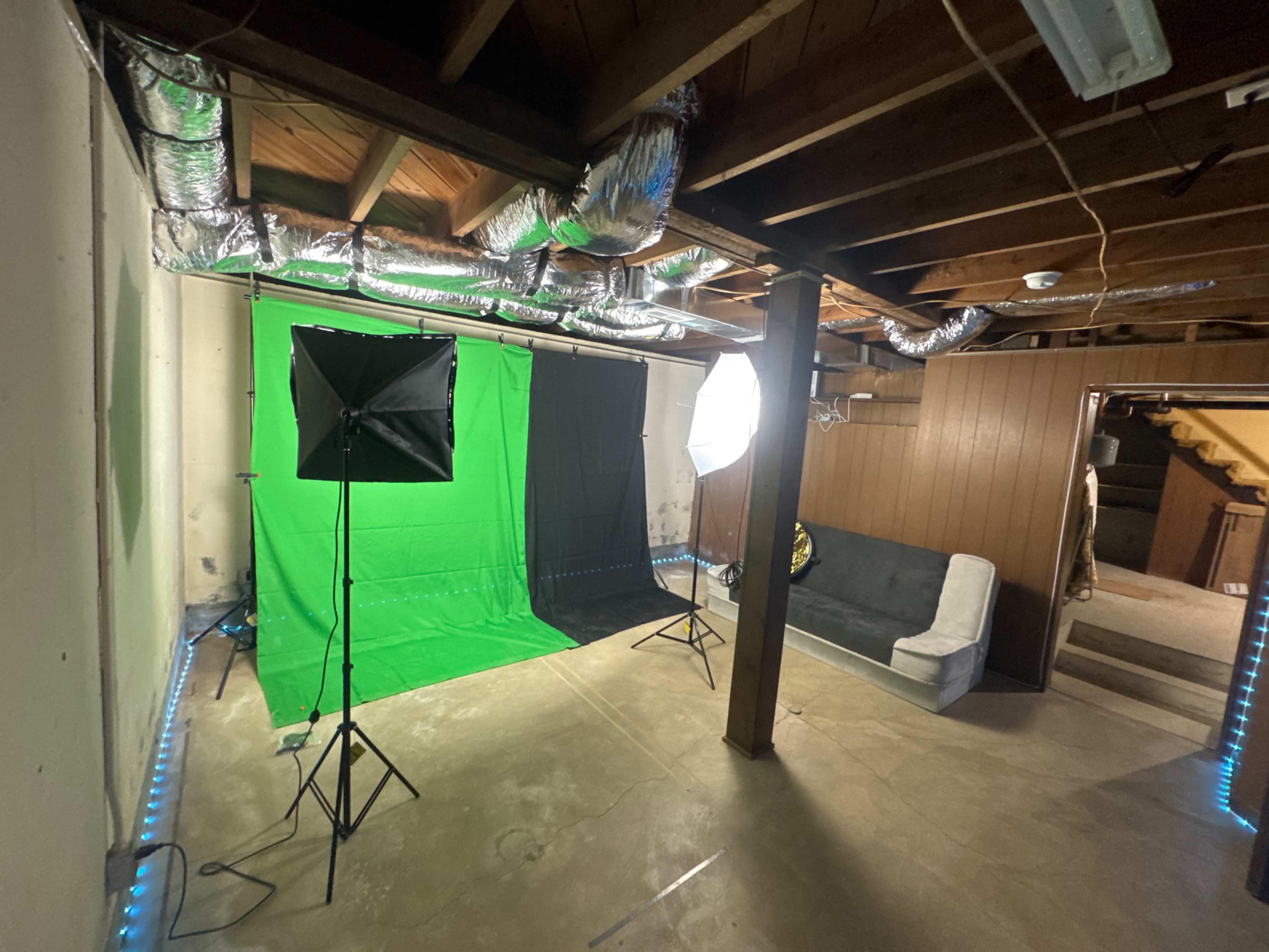 The image shows a basement studio setup with a green screen, a black backdrop, and lighting equipment, along with a couch against a wall.
