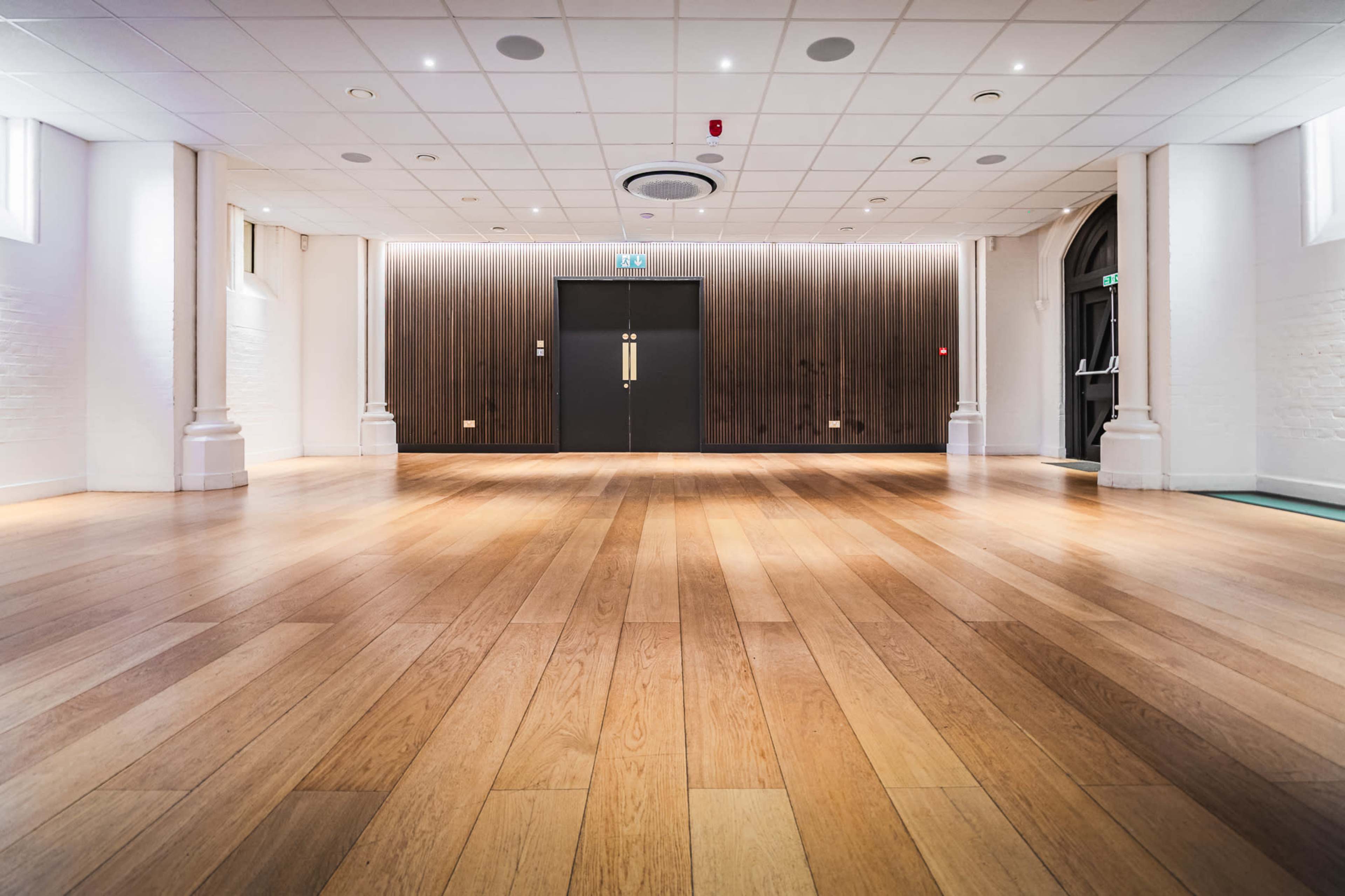 The image shows a spacious room with wooden flooring, white walls, and a dark-panelled wall at the far end featuring double doors.