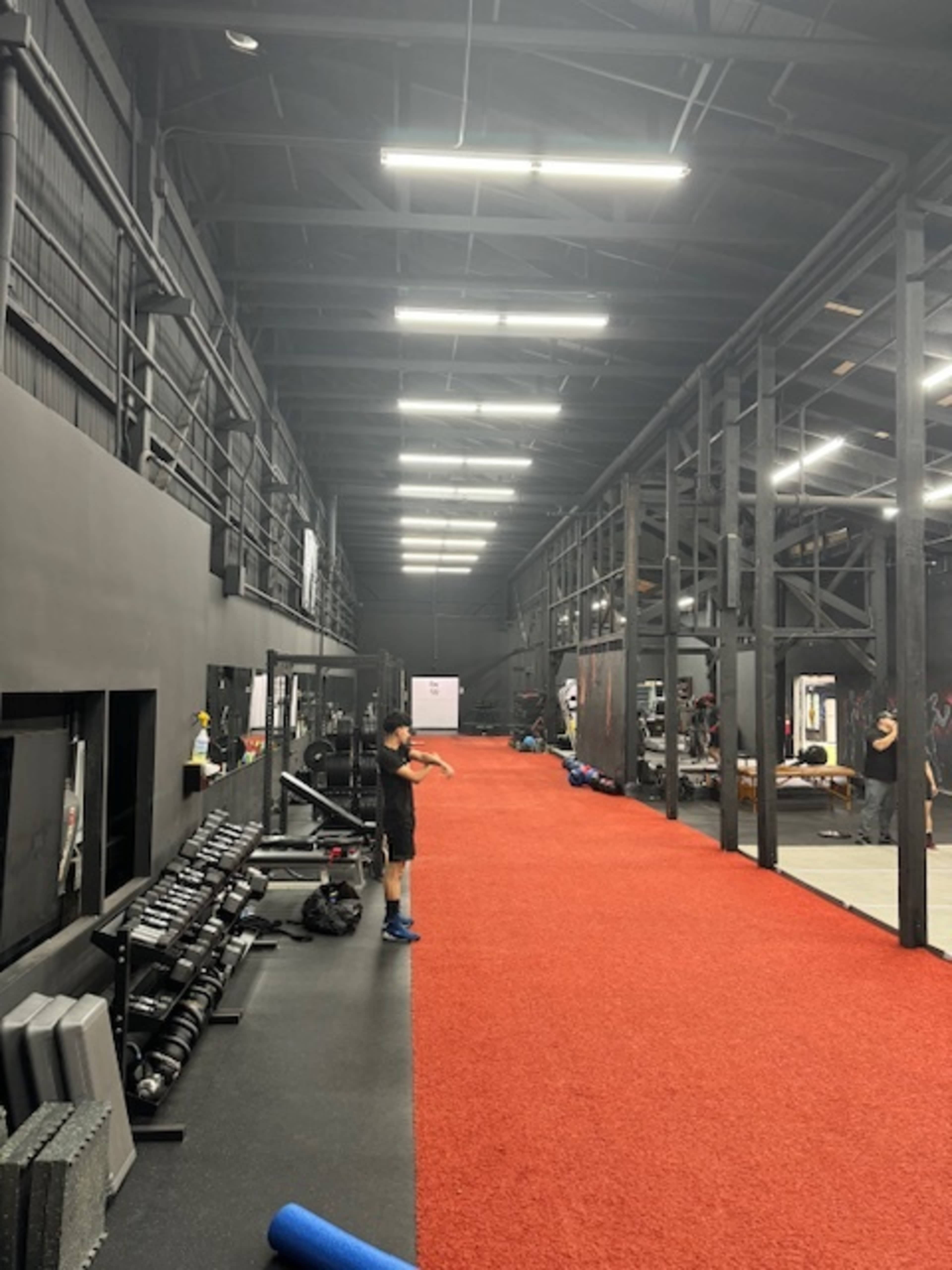 A large gym interior with a red carpeted pathway running through workout areas equipped with various exercise machines and weights.