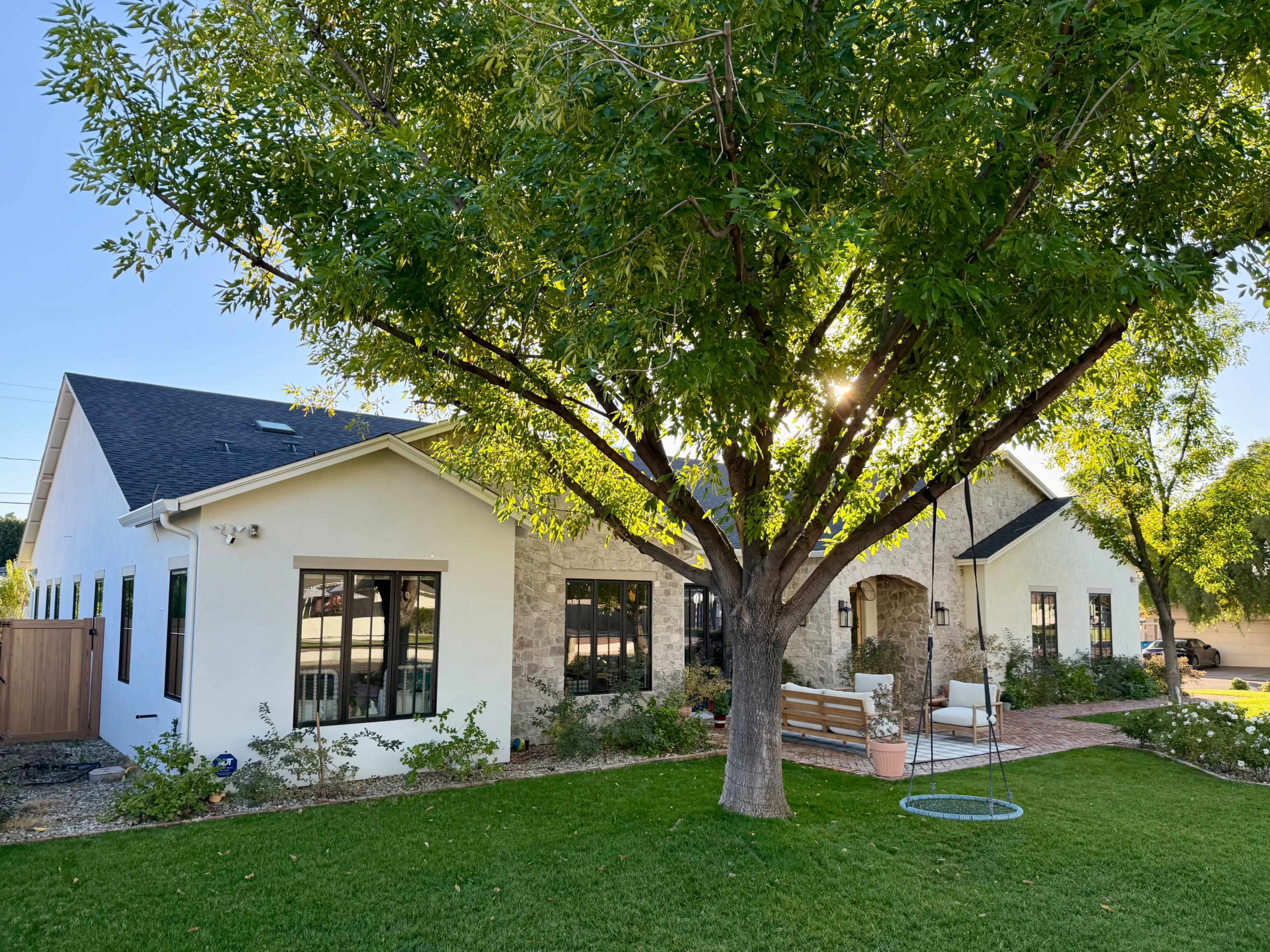 A white and stone house with large windows and a shaded porch sits next to a green lawn and a large tree.