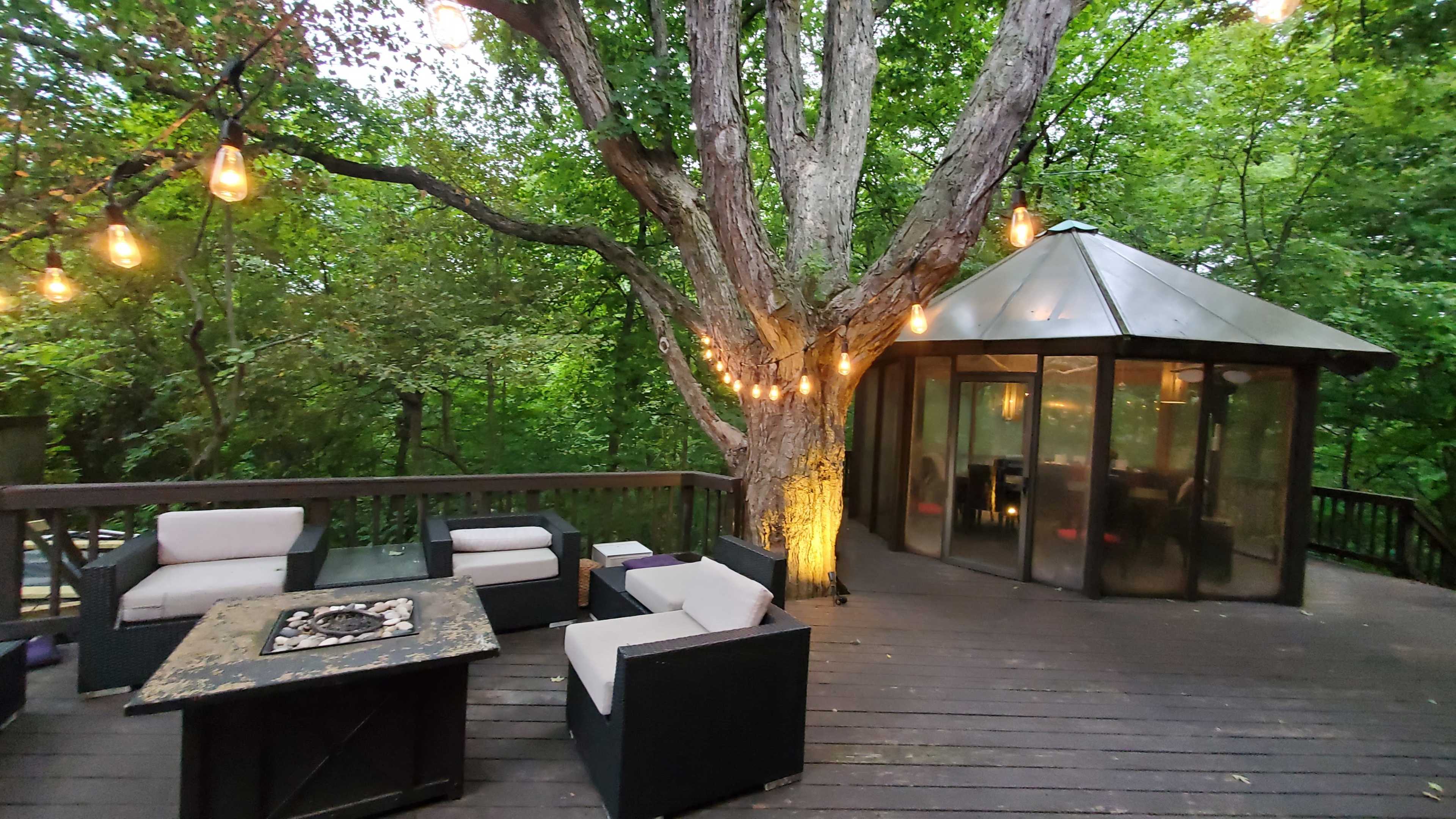 A wooden deck features comfortable seating, a fire pit, and a gazebo surrounded by trees illuminated with hanging lights.