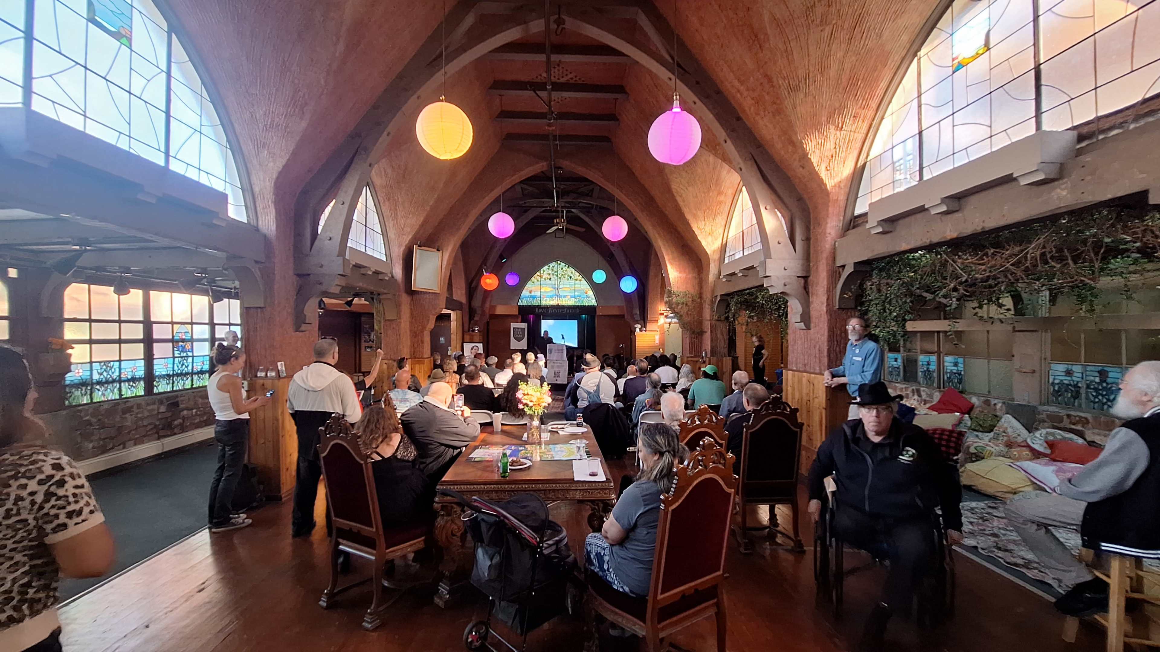 The image shows a large gathering in a rustic hall with wooden beams, colorful hanging lanterns, and attendees seated at tables and in chairs, while a presentation is displayed on a screen at one end.