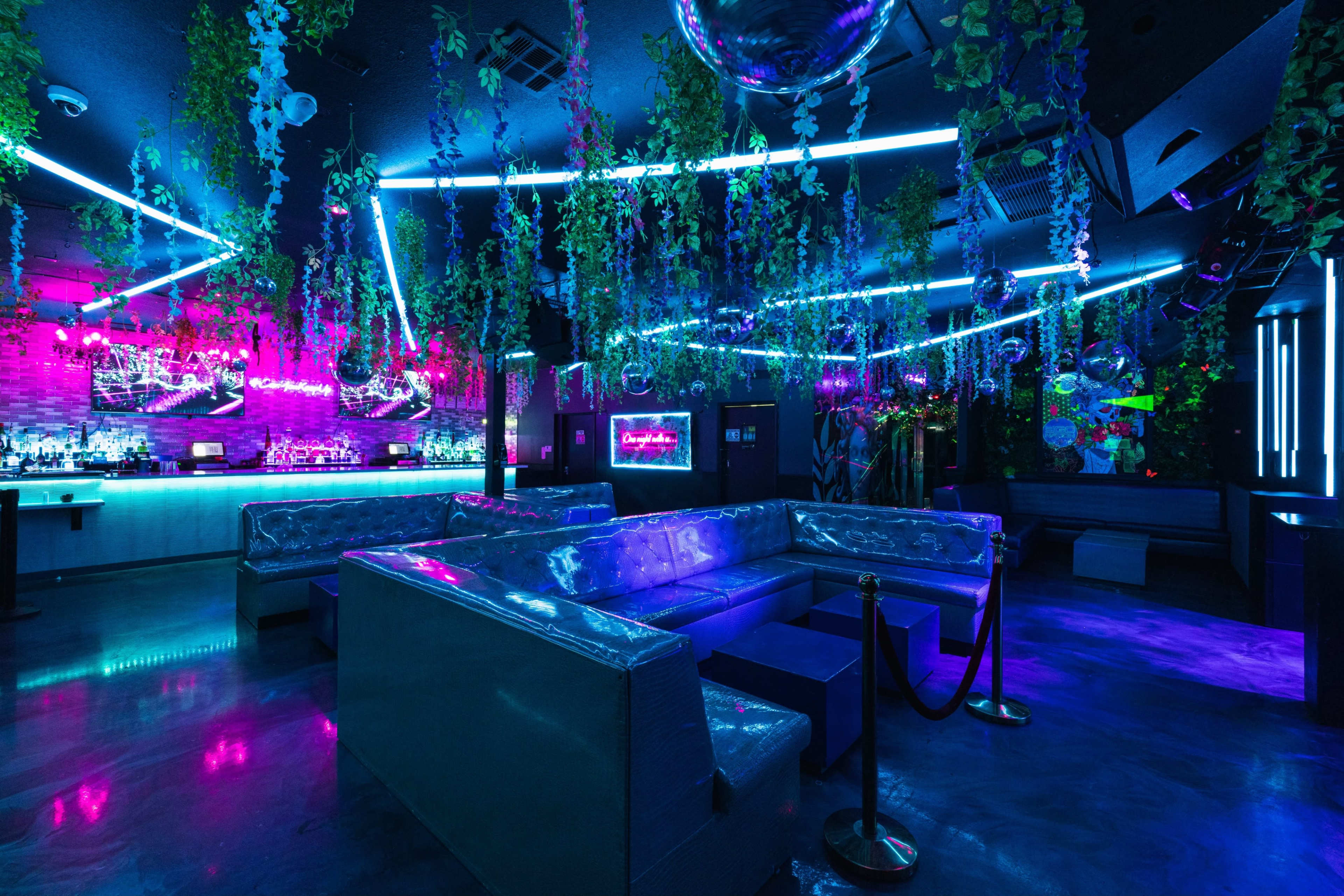 A brightly lit nightclub features neon lights, decorative plants, and seating areas arranged around a bar.