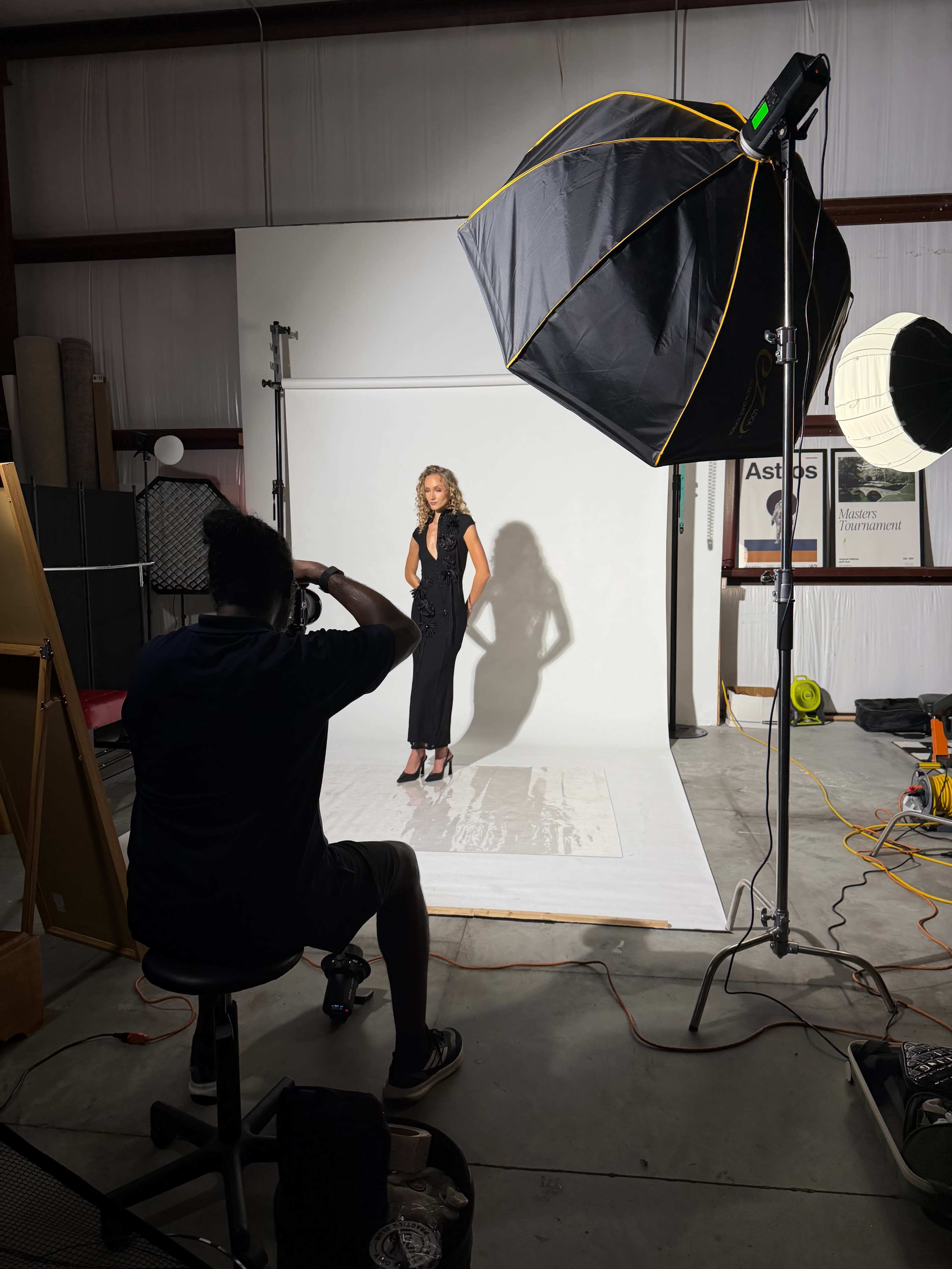 A photographer takes a portrait of a model against a white backdrop in a studio, using multiple lighting setups.