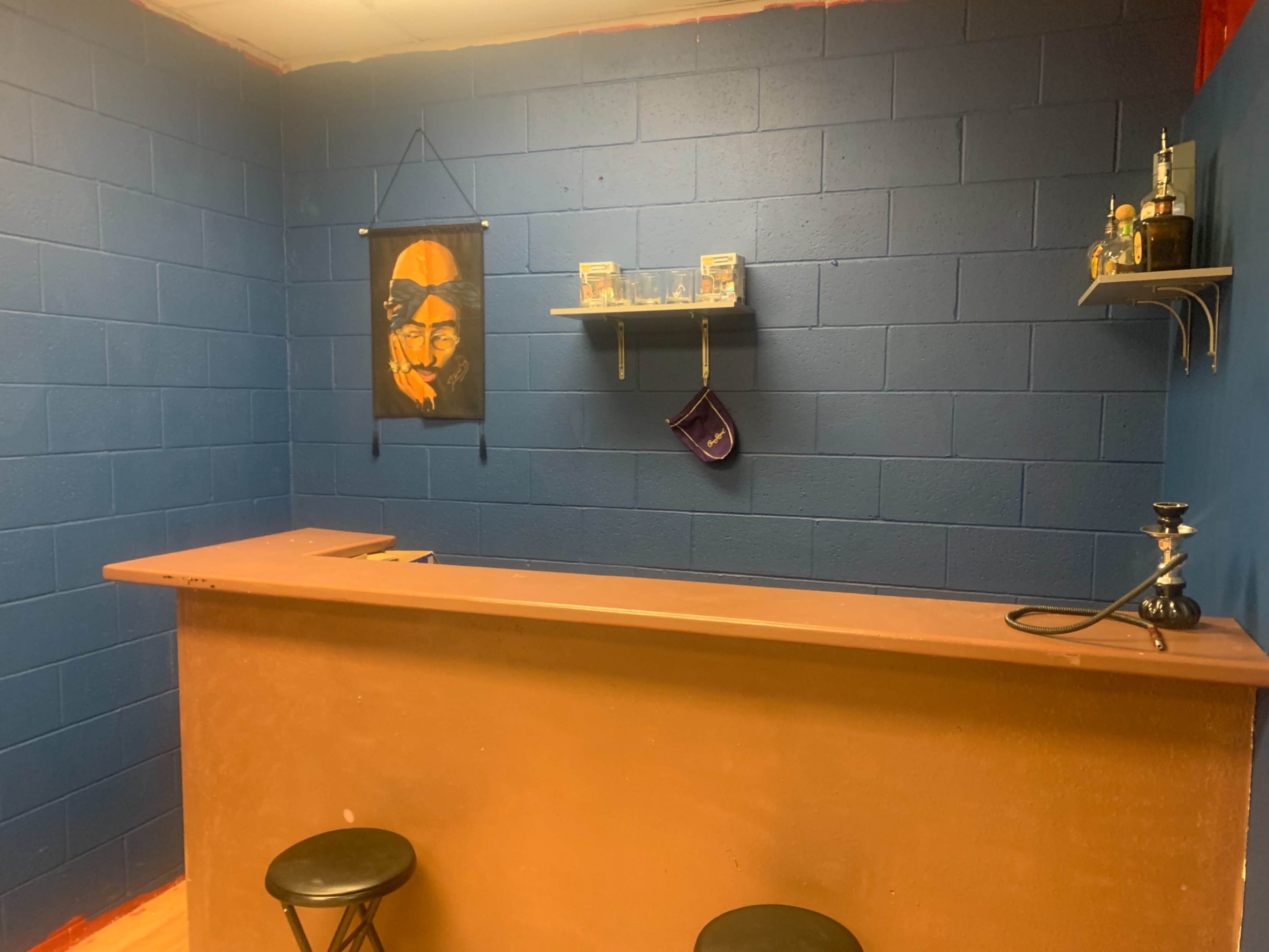 A small bar area with a wooden counter, two black stools, a blue wall, and a shelf displaying glassware and a hookah.