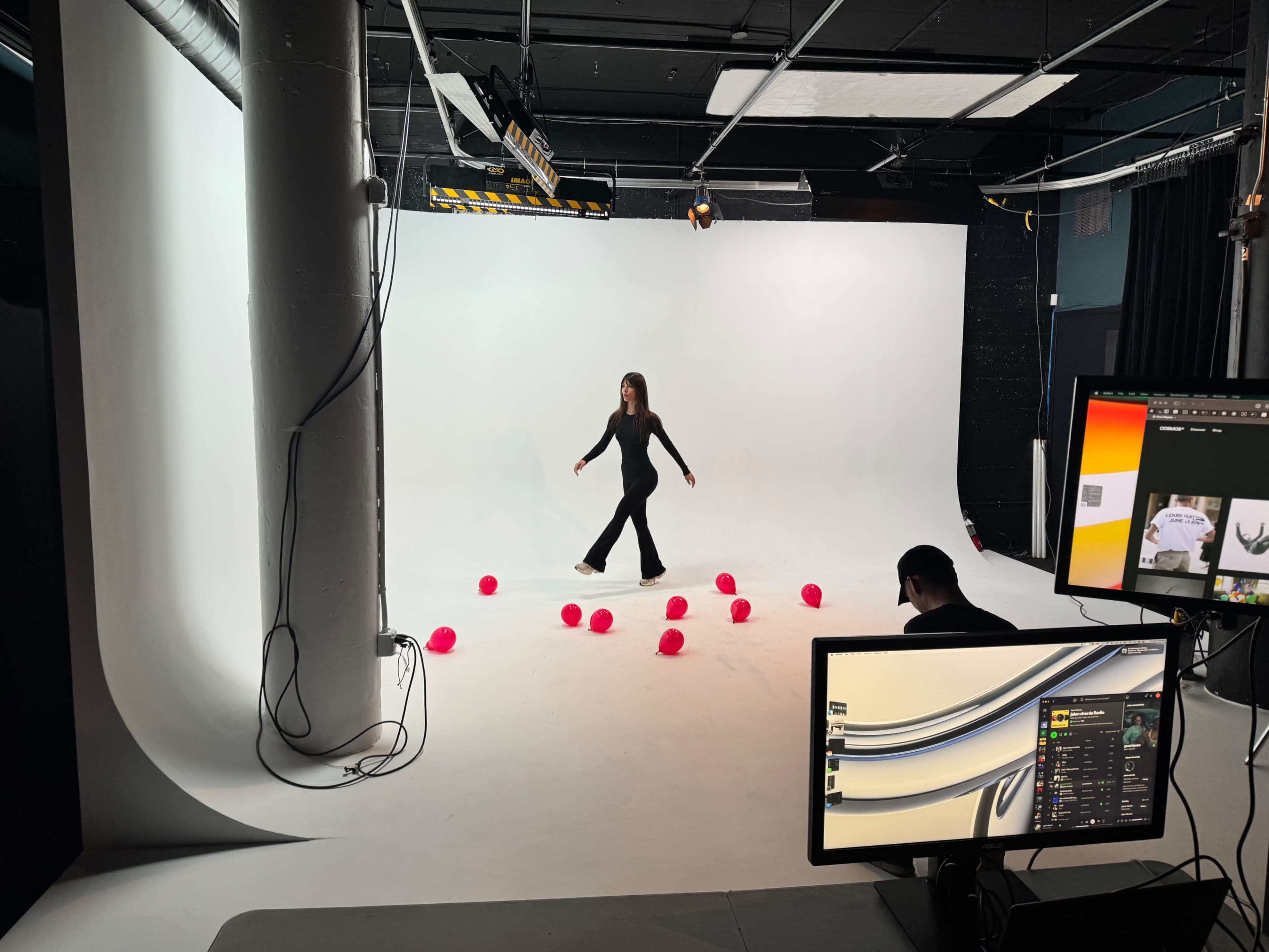 A woman dressed in black is walking through a studio surrounded by bright pink balls on a white backdrop, while a man operates a computer in the foreground.