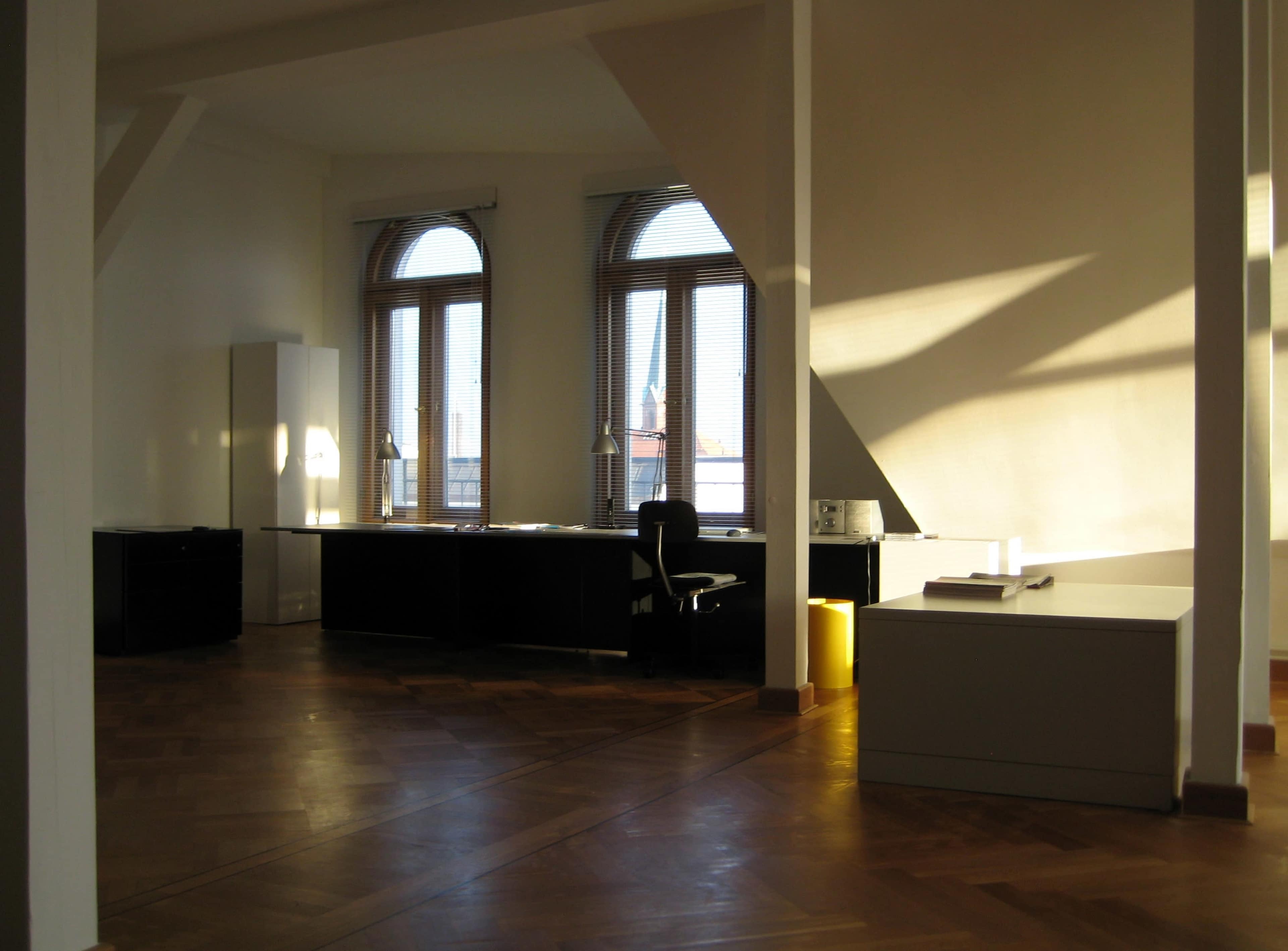 The image shows a bright, spacious office with large windows, wooden floors, and minimal furniture including a desk and filing cabinet.