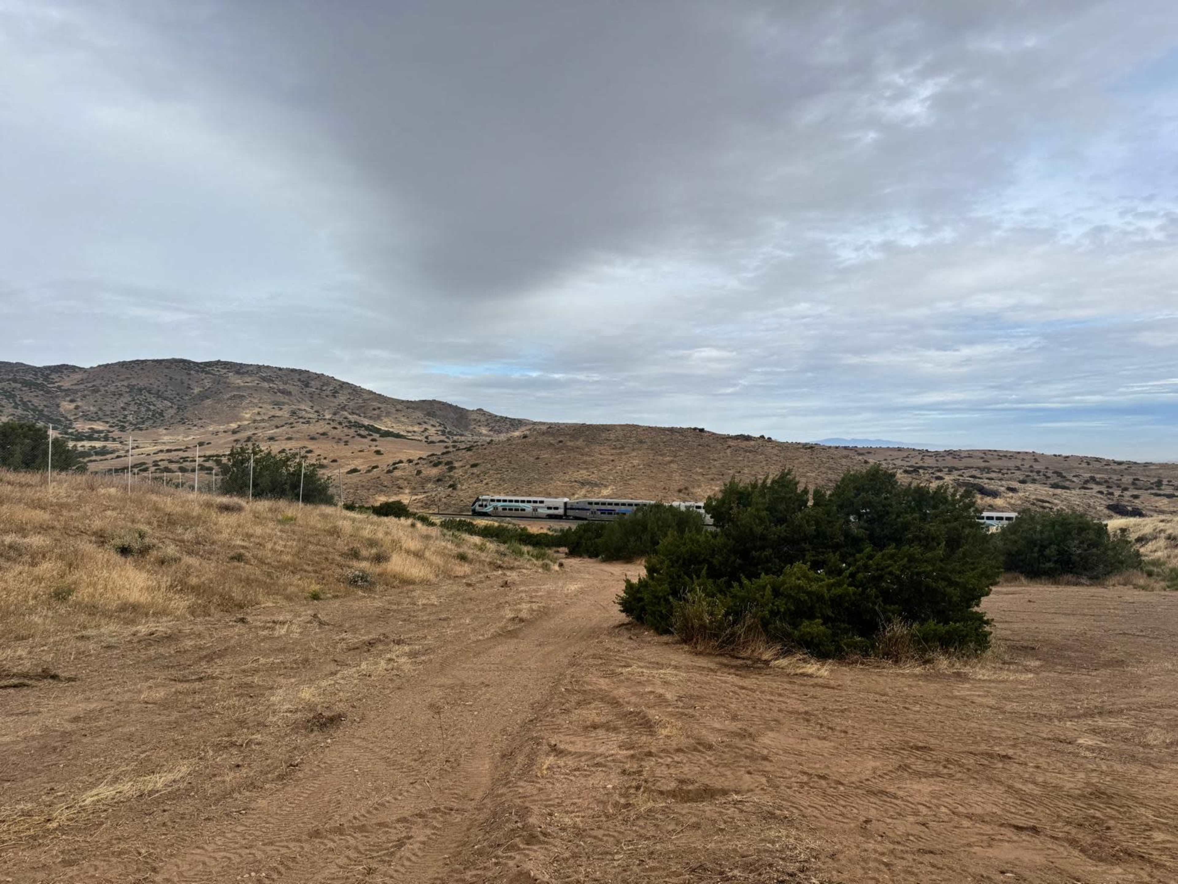 A train travels along tracks in a dry, hilly landscape with sparse vegetation under a cloudy sky.