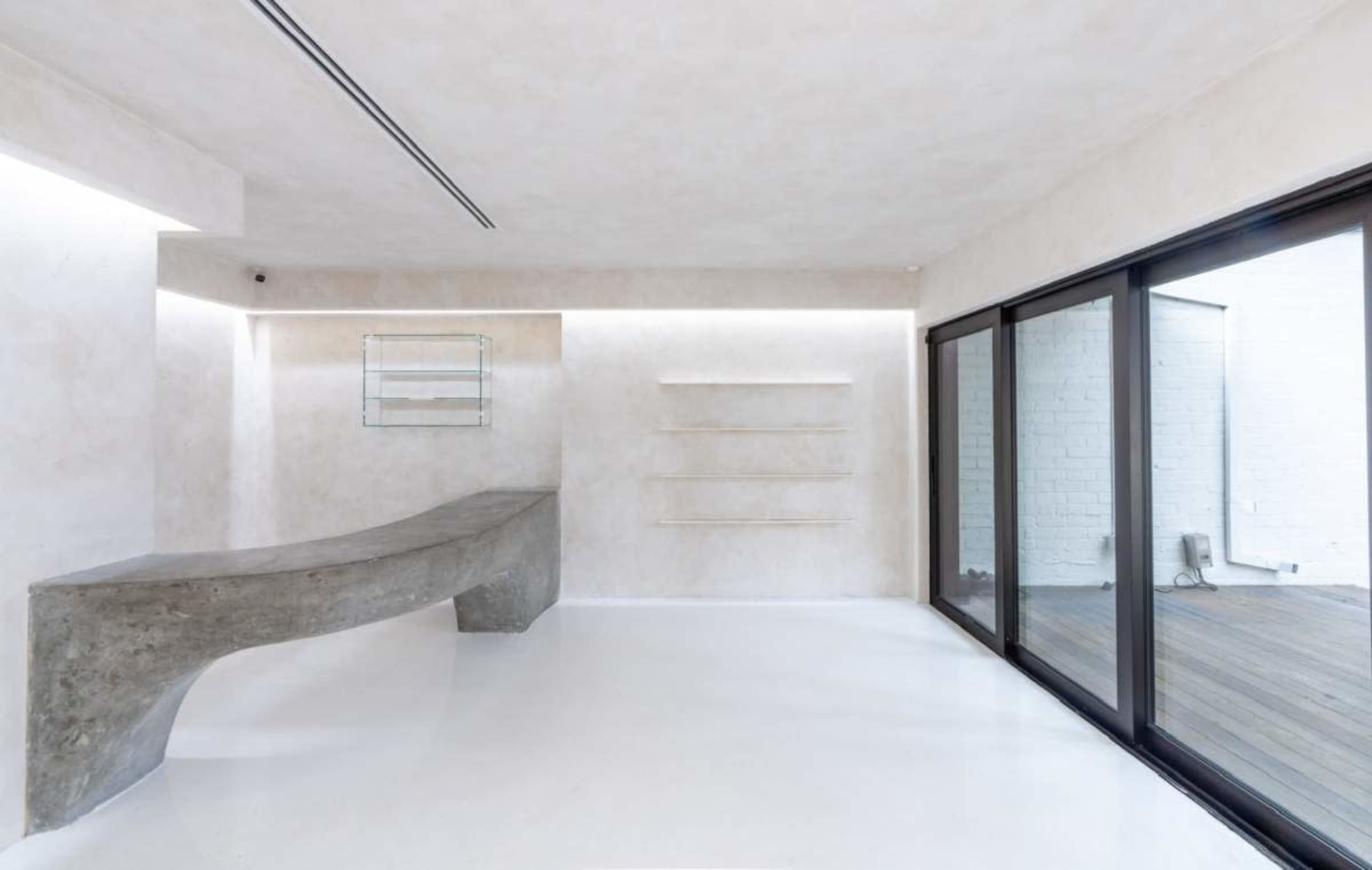 The image shows a minimalist interior space with a curved concrete reception desk, white walls, and large glass sliding doors opening to a wooden deck.