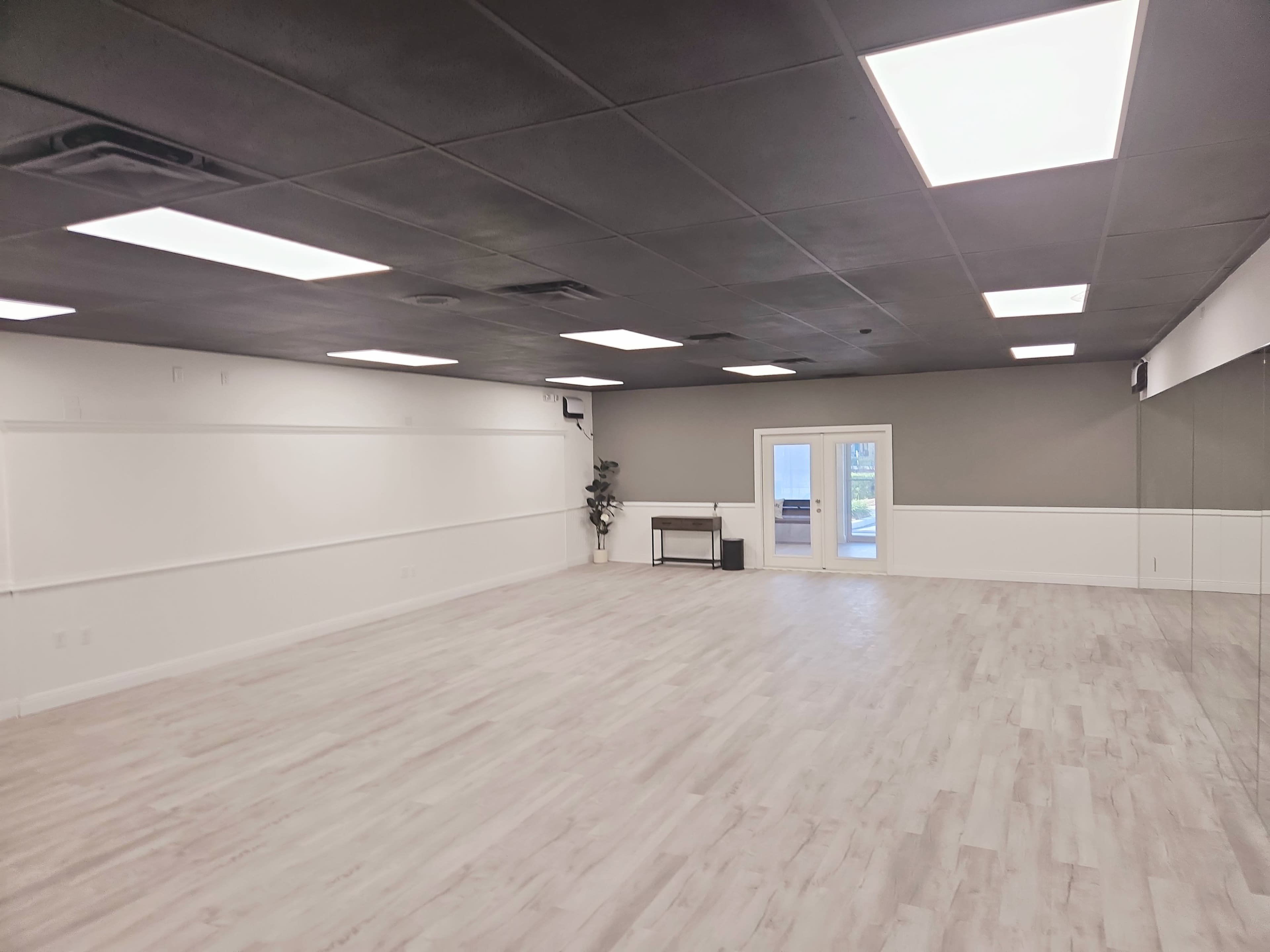 The image shows a spacious, empty room with light-colored flooring, white walls, and several overhead lights, featuring a door opening to an outdoor area.