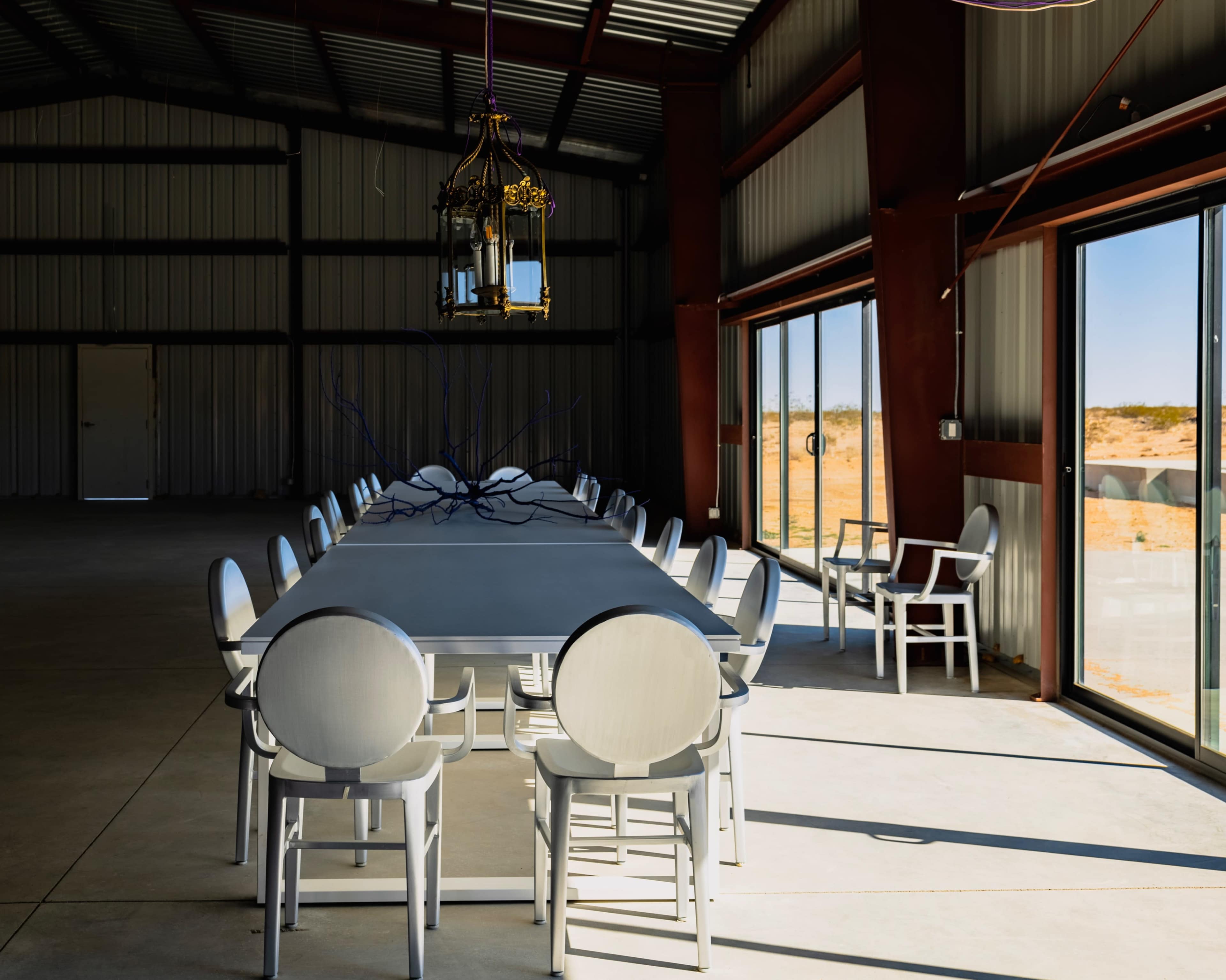 A long, white dining table with matching chairs is positioned in a spacious, industrial-style room with large windows revealing a desert landscape outside.