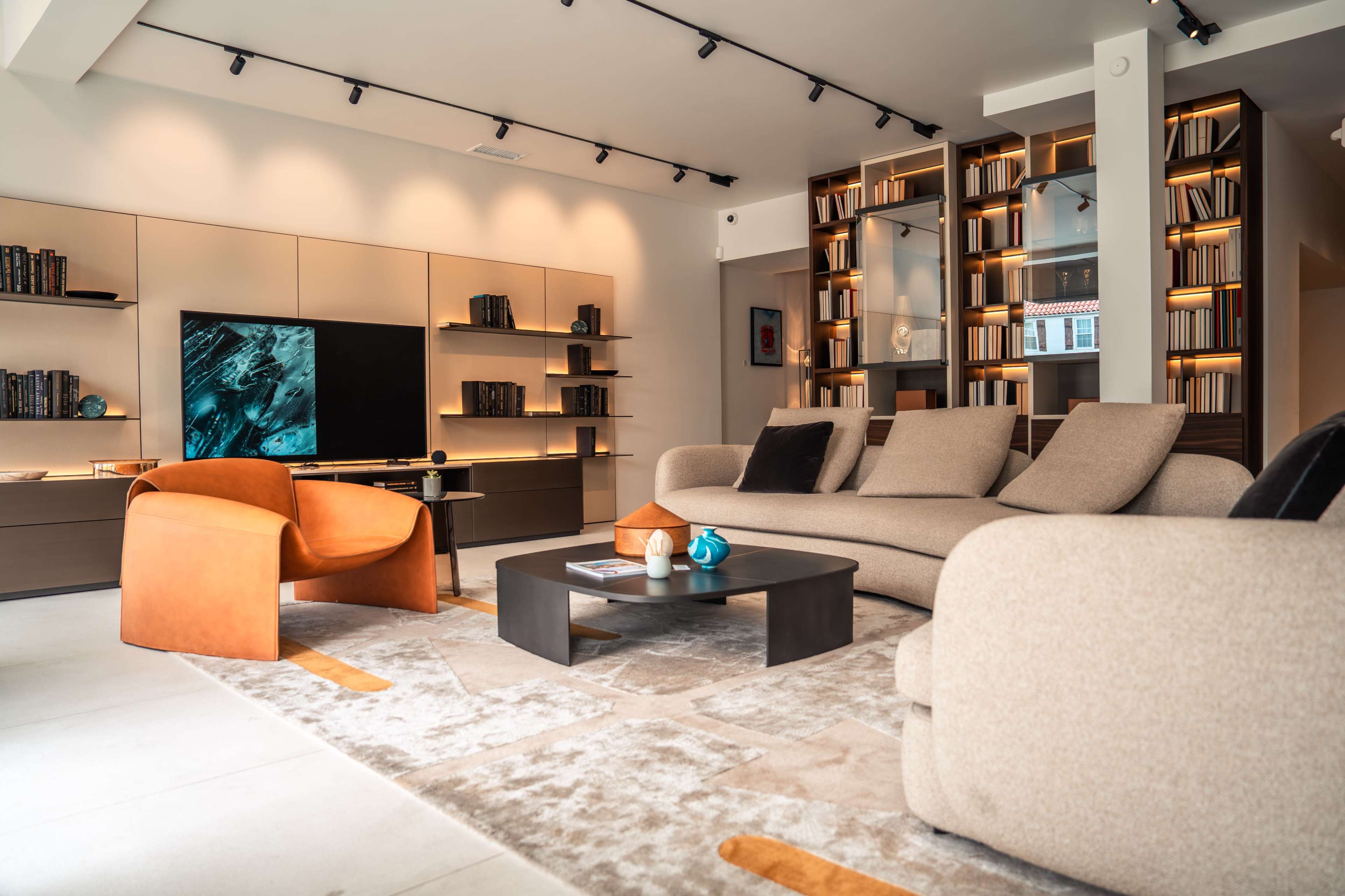 The image depicts a modern living room featuring a beige sofa, an orange armchair, a black coffee table, a television mounted on a wall unit, and bookshelves illuminated with warm lighting.