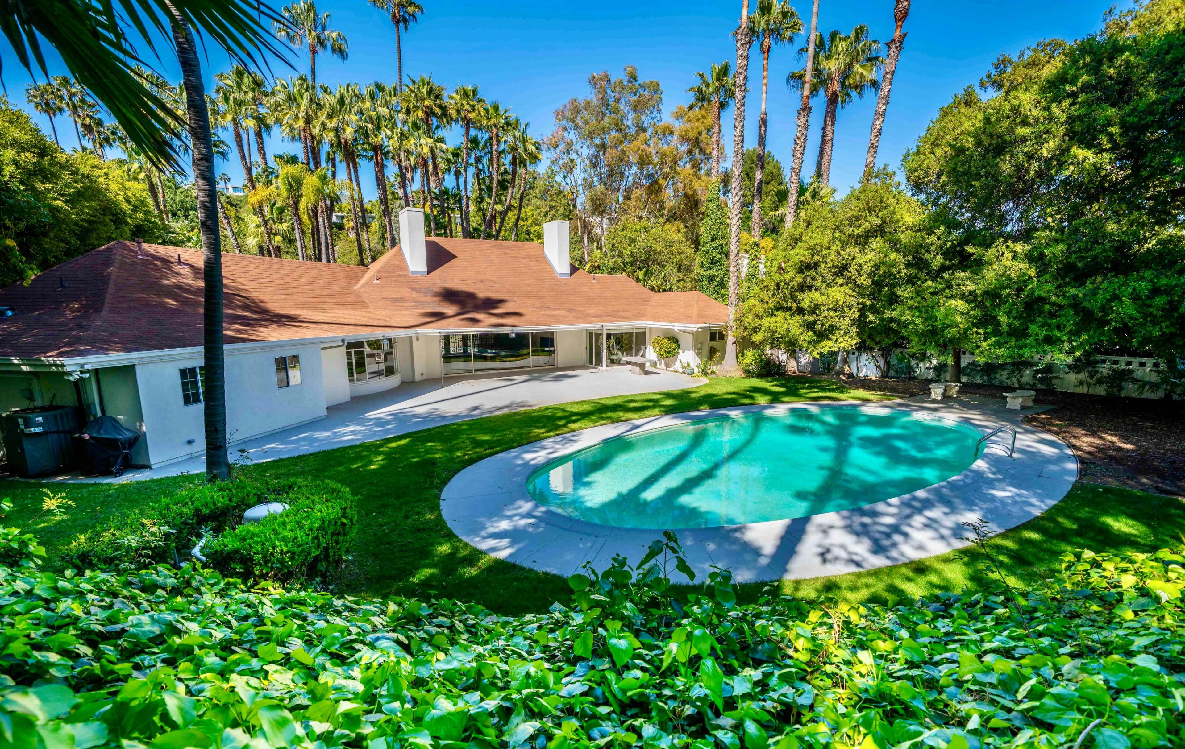 The image shows a backyard with a swimming pool surrounded by lush greenery and palm trees, adjacent to a single-story house.