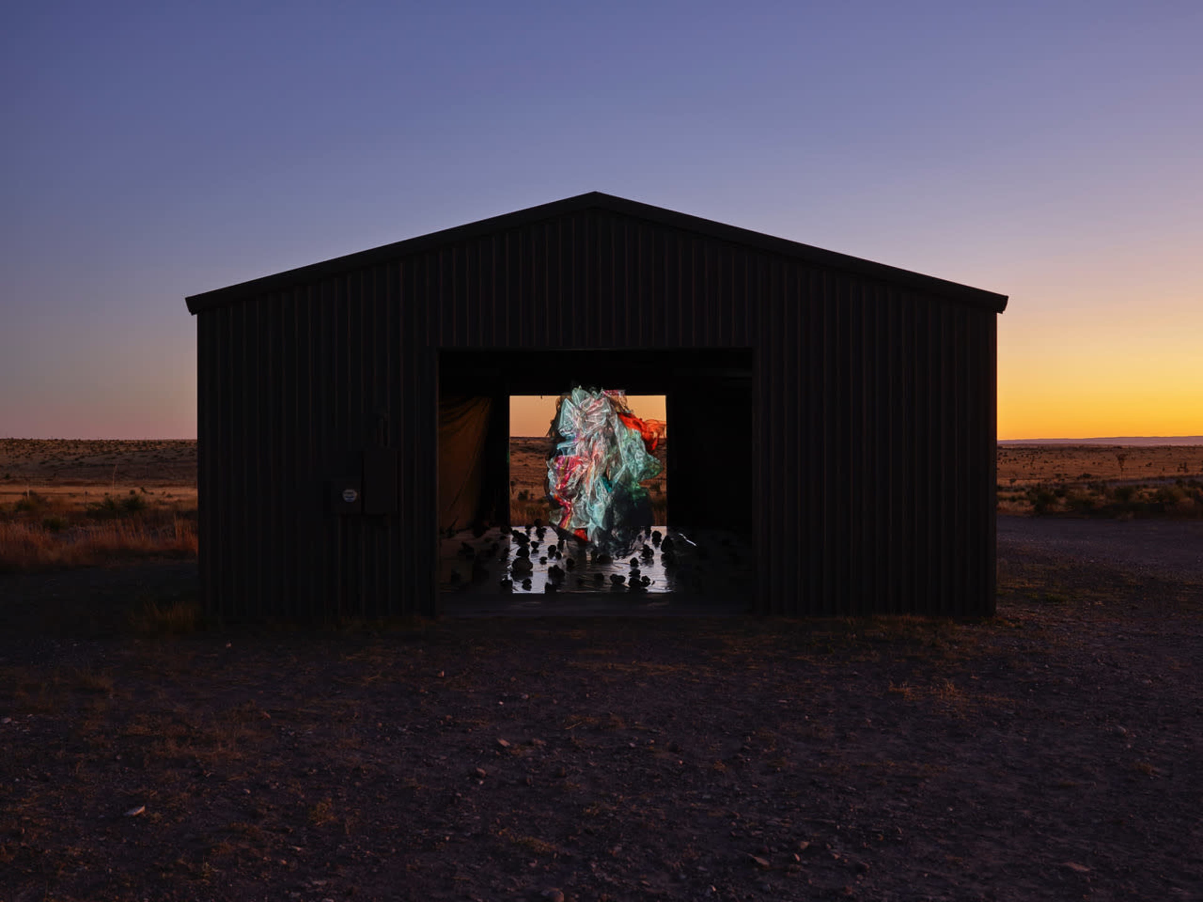 A large, abstract sculpture is displayed inside a dark building, illuminated by the soft glow of dusk outside.