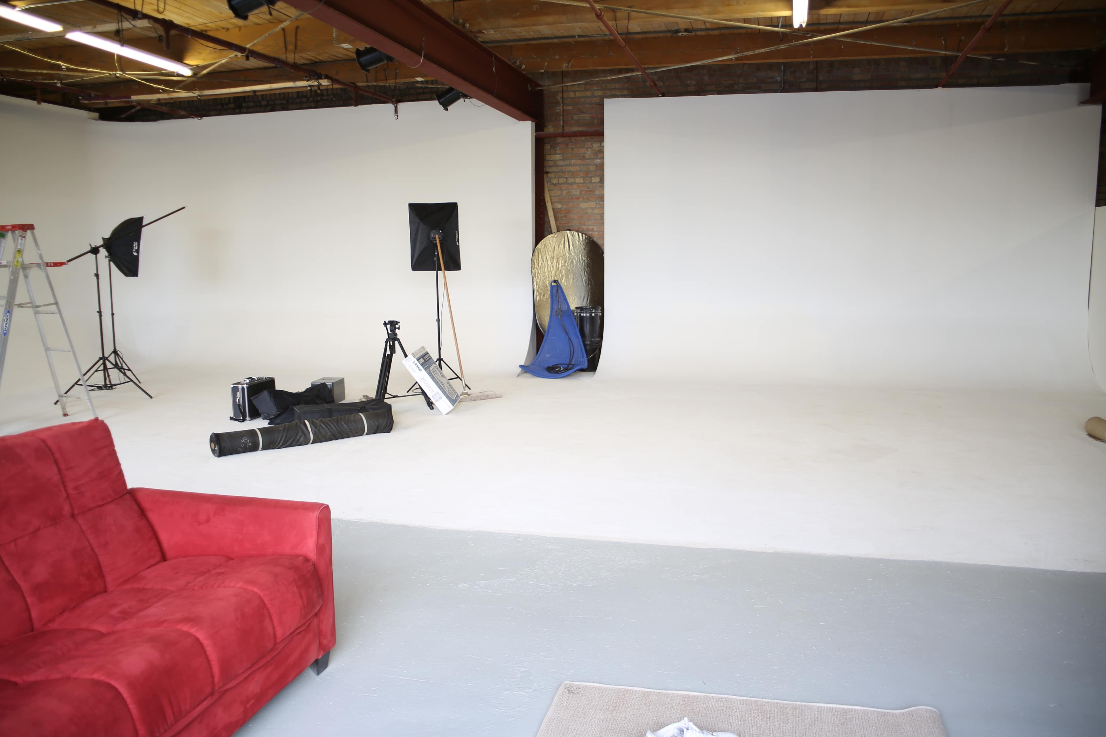 A spacious photography studio featuring a red couch, a ladder, and various equipment set against blank white walls.